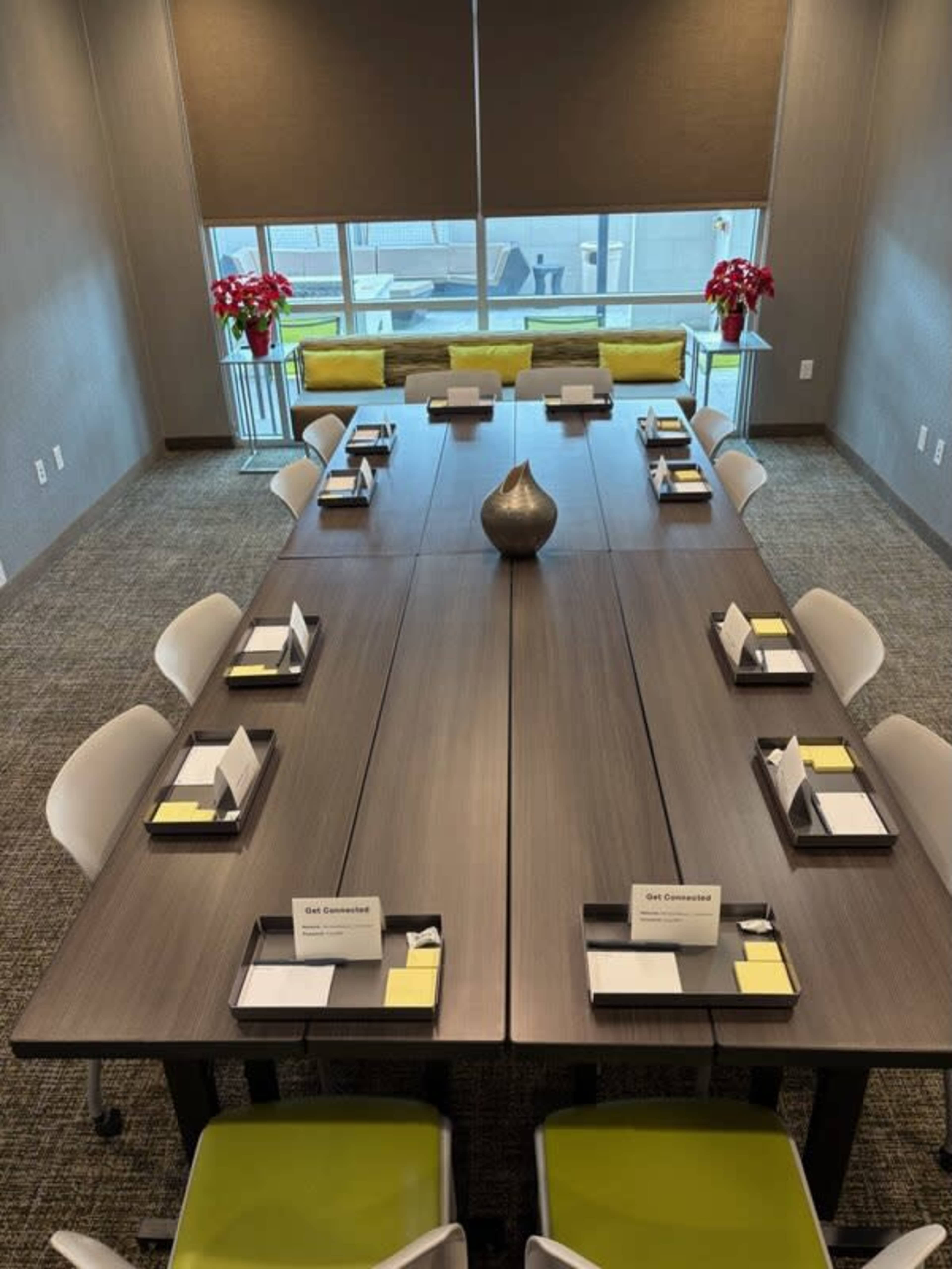 A long conference table sits in a meeting room, surrounded by chairs, with place settings and yellow notepads at each spot, and large windows allowing natural light to enter the space.