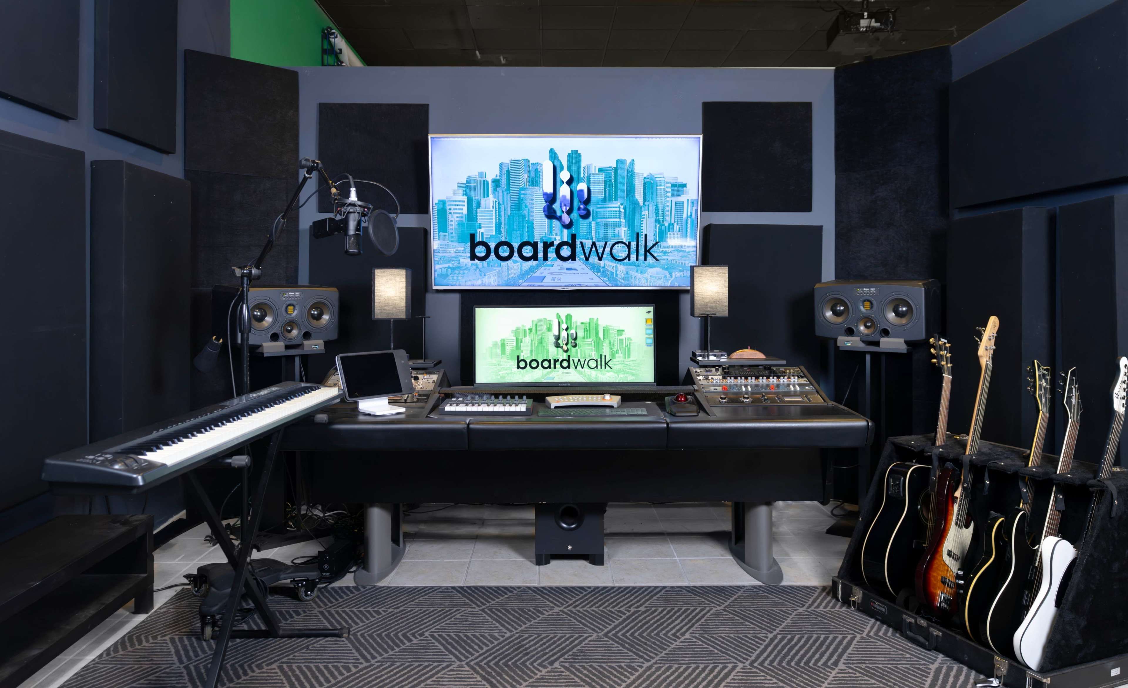 A music studio with a large mixing console, two computer monitors displaying the logo "boardwalk," a keyboard, speakers, and several electric guitars on a rack.