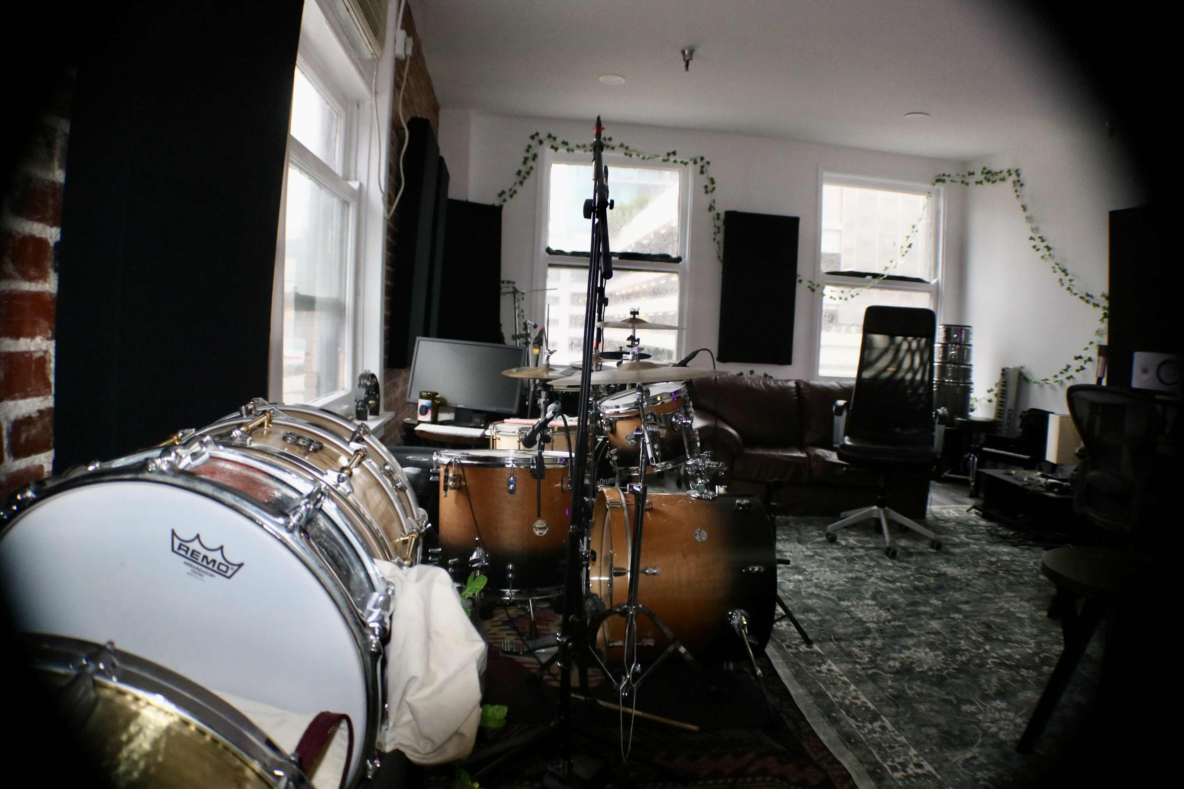 The image shows a music studio with a drum set, a desk chair, and various sound equipment arranged in a room featuring brick walls and a patterned rug.