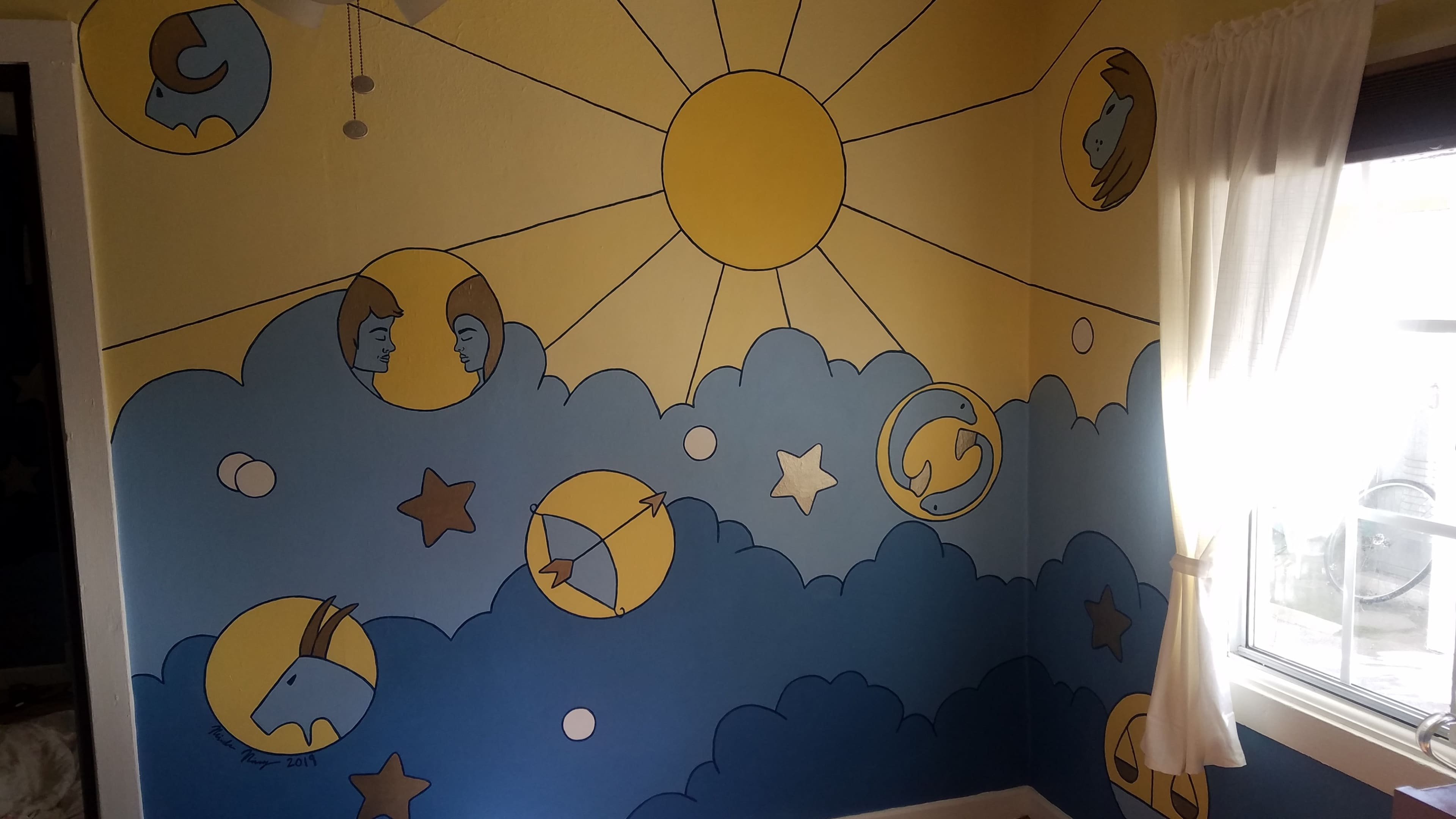 The image shows a mural on a wall featuring a sun, clouds, and various celestial figures and shapes in a blue and yellow color scheme.