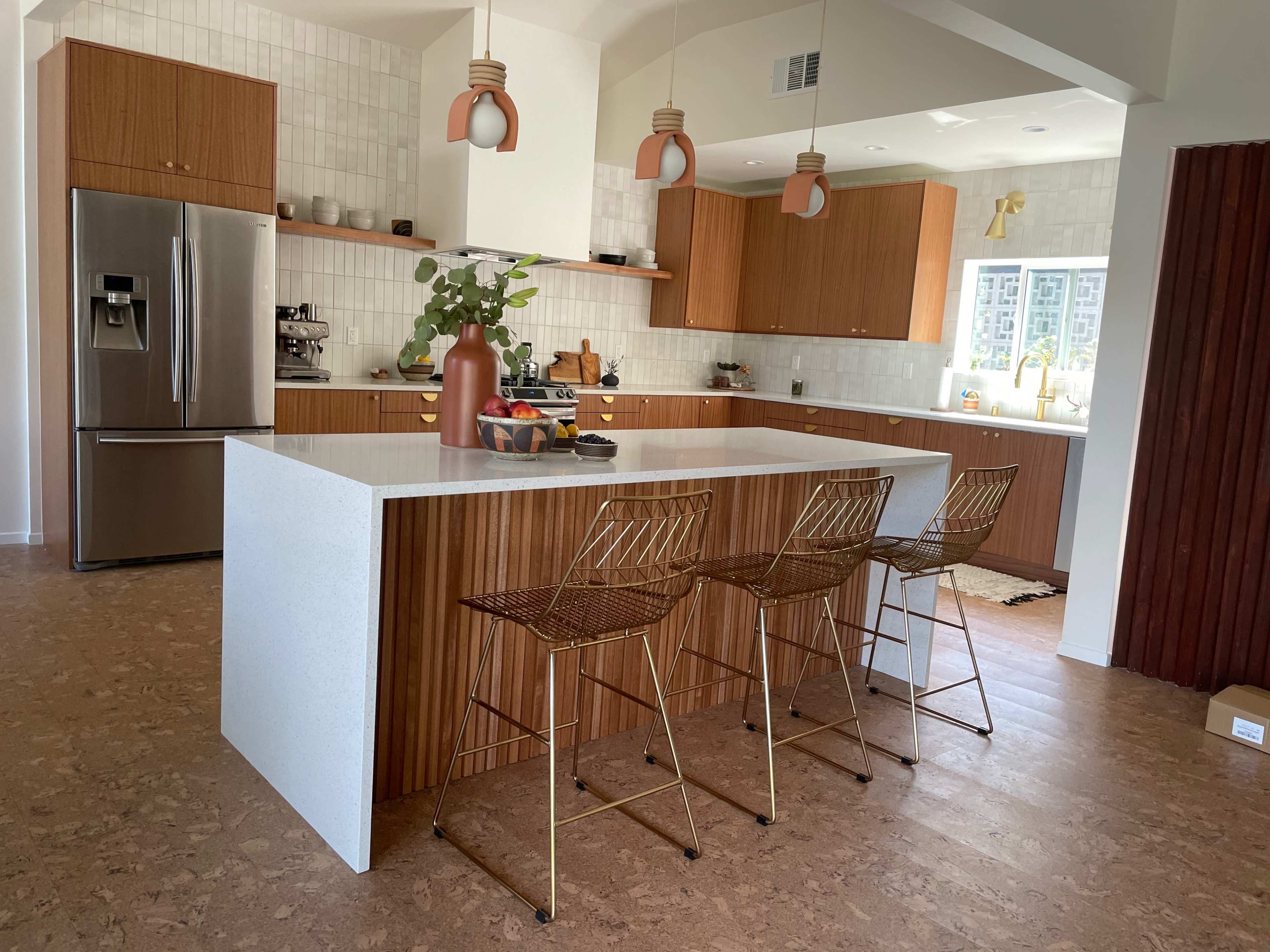 A modern kitchen features a large white island with three metal bar stools, wooden cabinetry, and light fixtures hanging above.