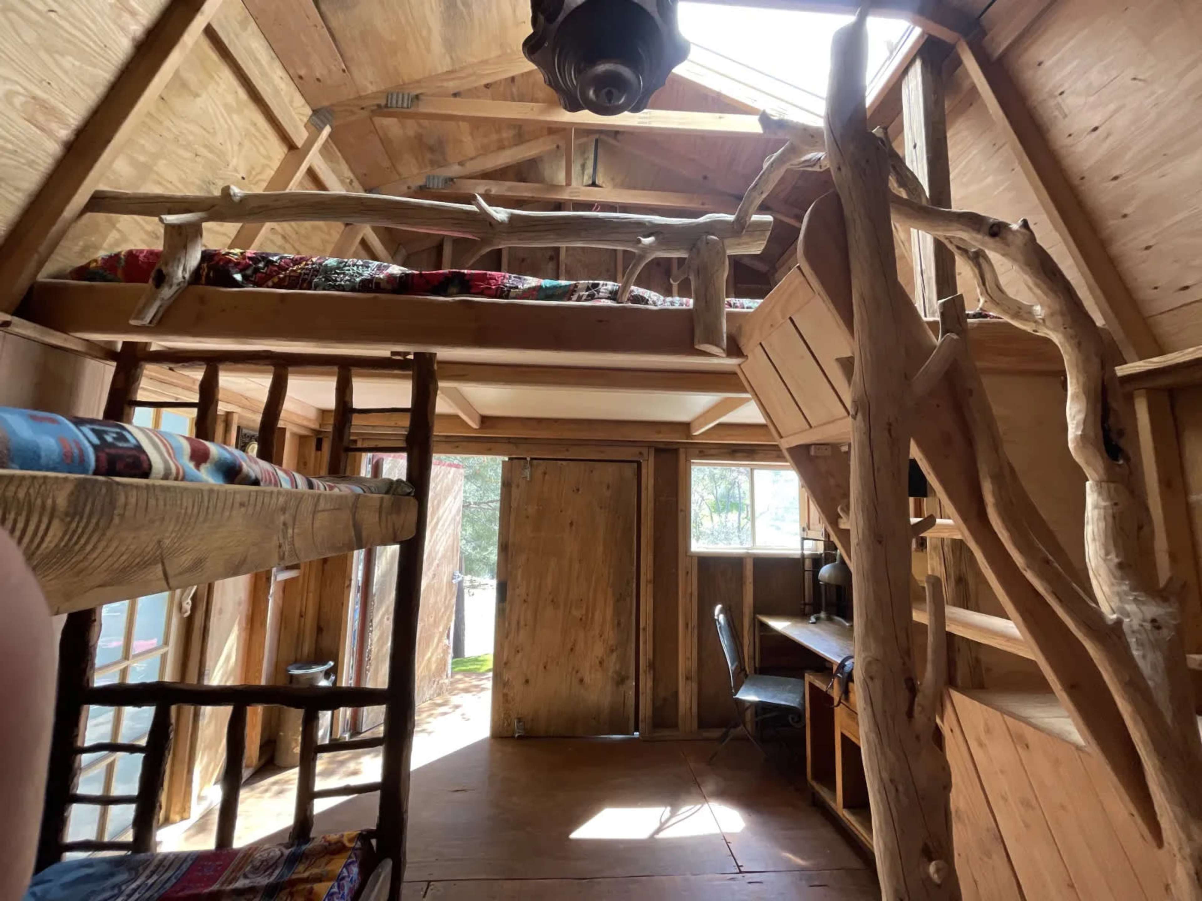 The interior of a wooden cabin features a lofted bed, a lower bunk bed, and a small desk area near a large window.