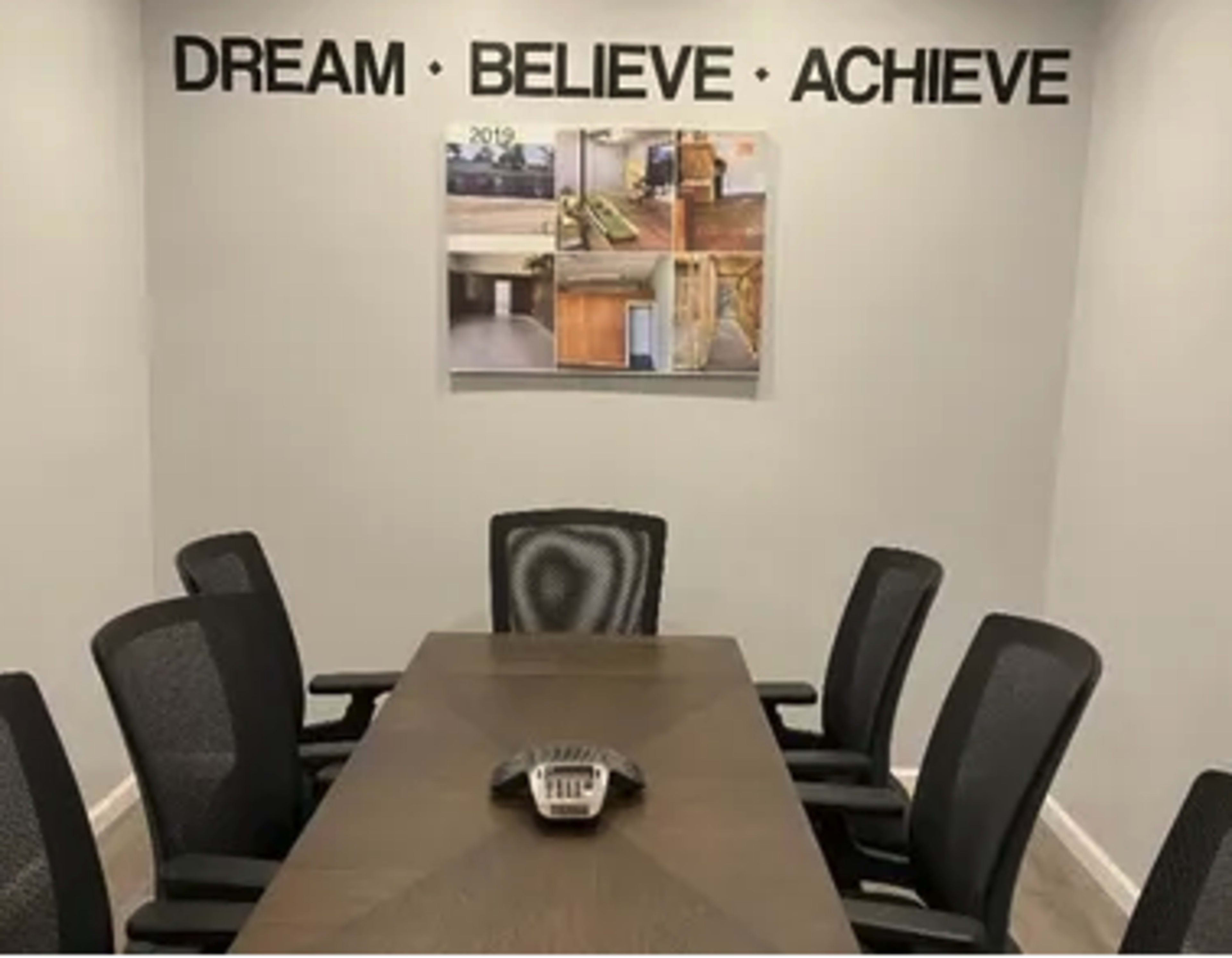 A conference room features a long table surrounded by black mesh chairs, with the words "DREAM BELIEVE ACHIEVE" displayed on the wall above a framed photo.