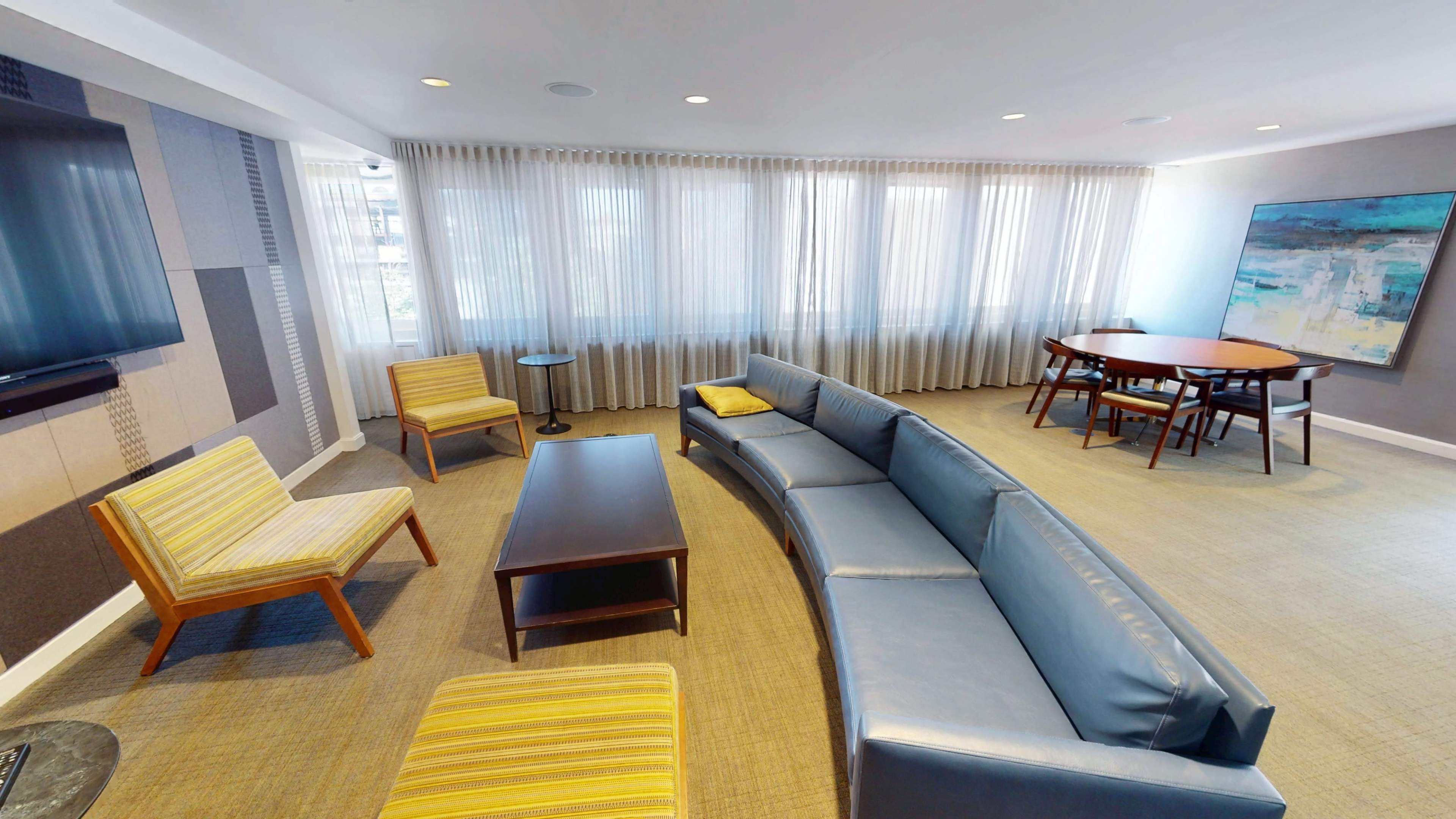 The image shows a modern lounge area with a curved sofa, two armchairs, a coffee table, and a dining table surrounded by large windows with sheer curtains.