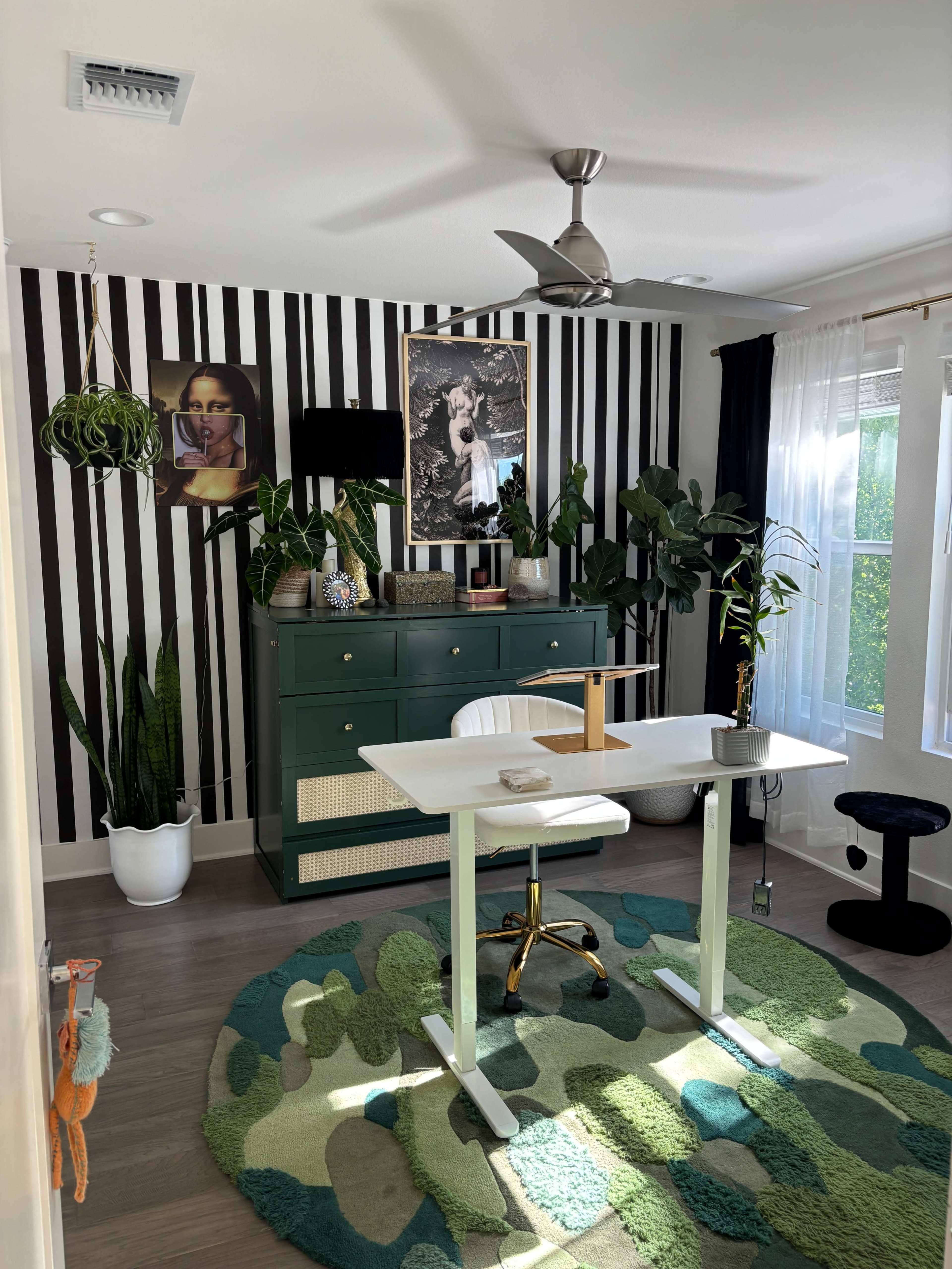 The image shows a modern home office with a white standing desk, a green rug, indoor plants, and black-and-white striped walls decorated with framed artwork.