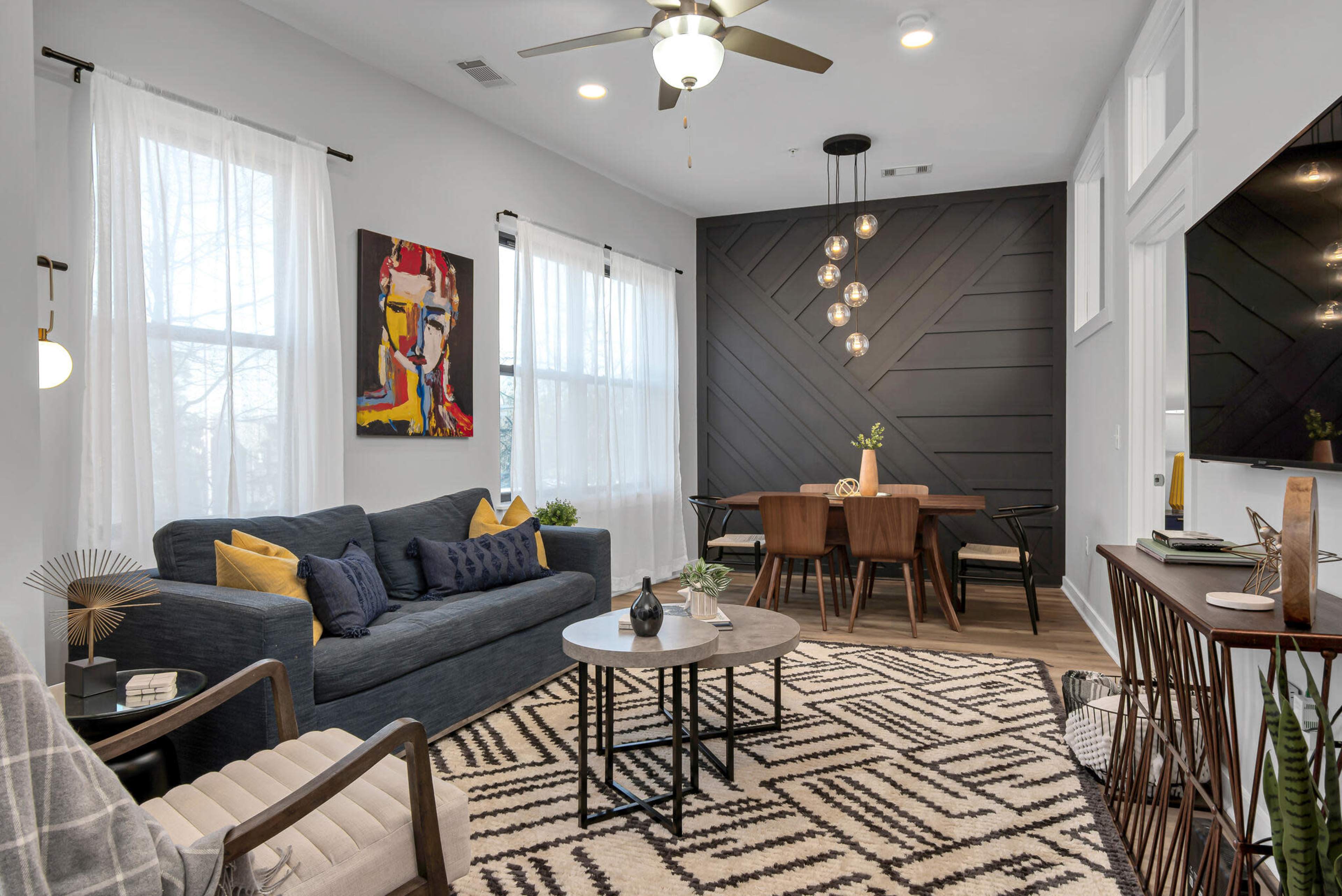 The image depicts a modern living room with a blue sofa, a round coffee table, and a dining area featuring a wooden table and chairs, all set against a backdrop of large windows and stylish wall designs.