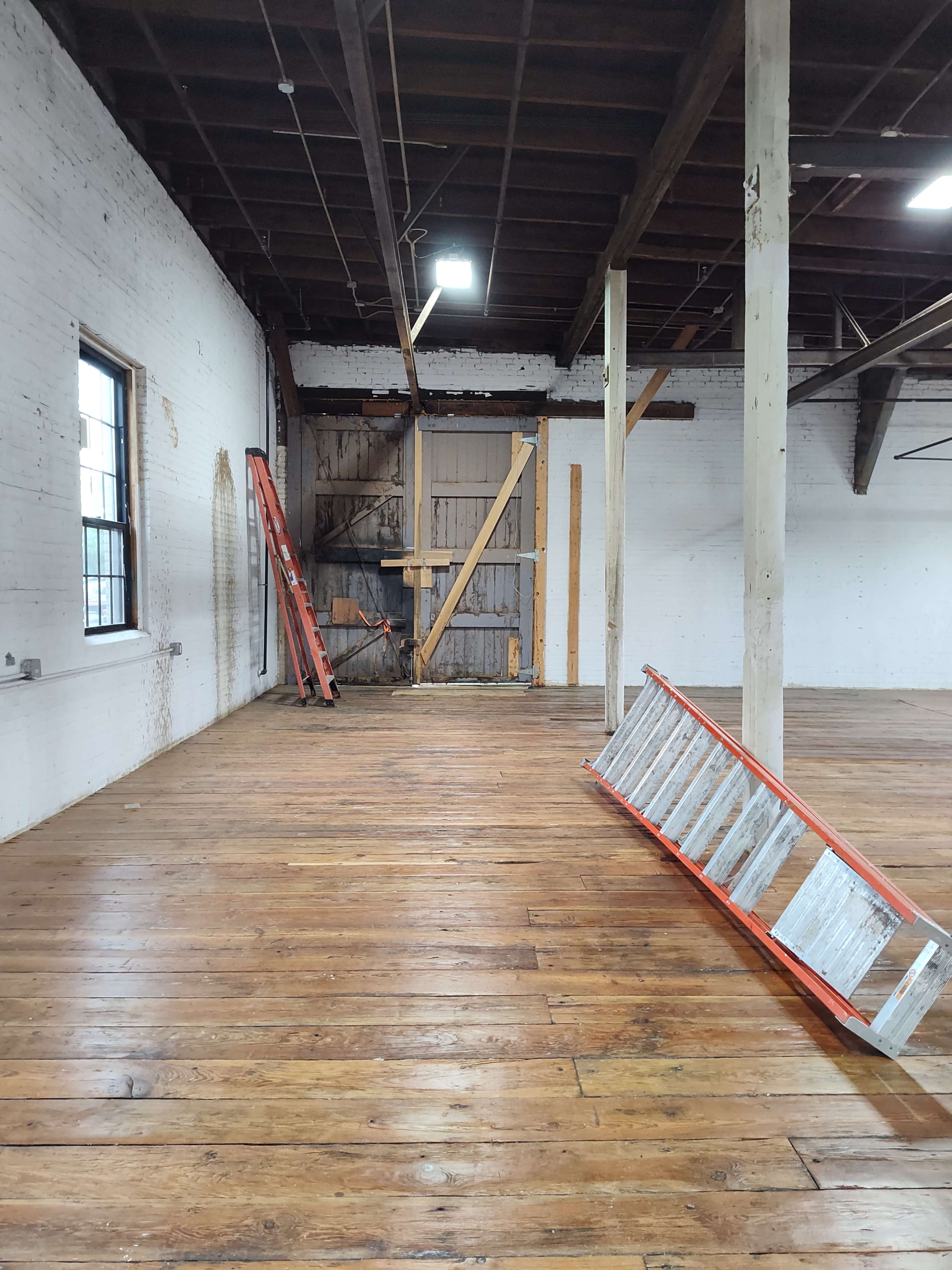 An empty industrial room features wooden flooring, exposed beams, and a partially open door with a ladder and a leaning extension ladder against the wall.
