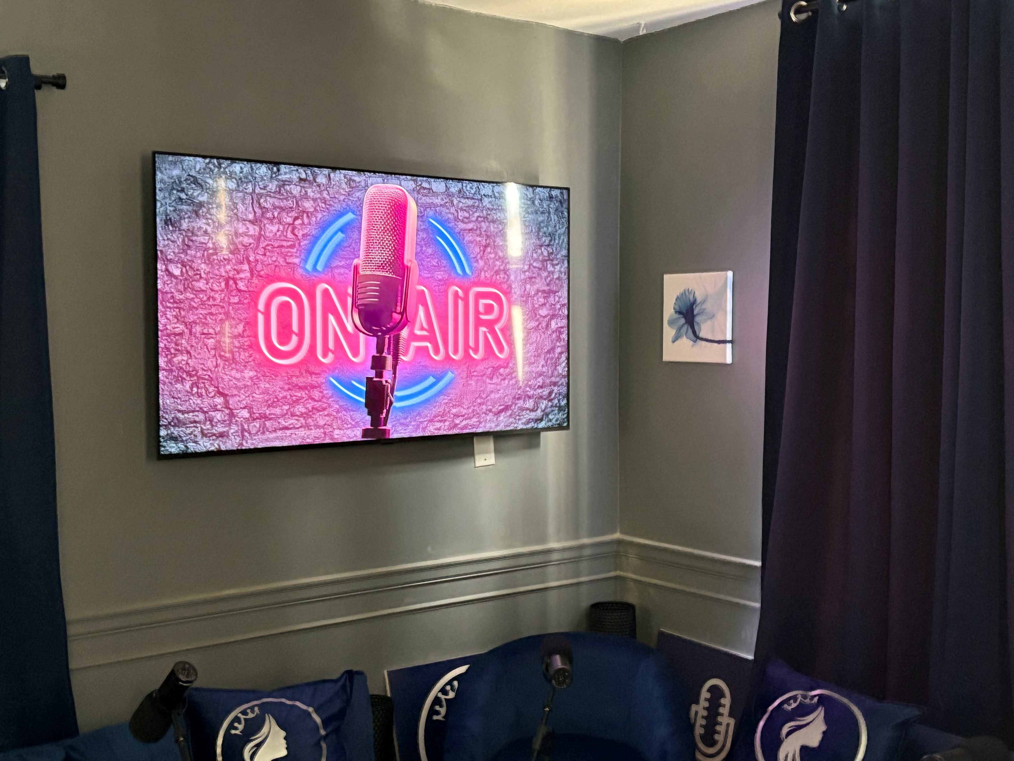 The image shows a wall-mounted television displaying an "ON AIR" neon sign, surrounded by a cozy setup with blue chairs and curtains.
