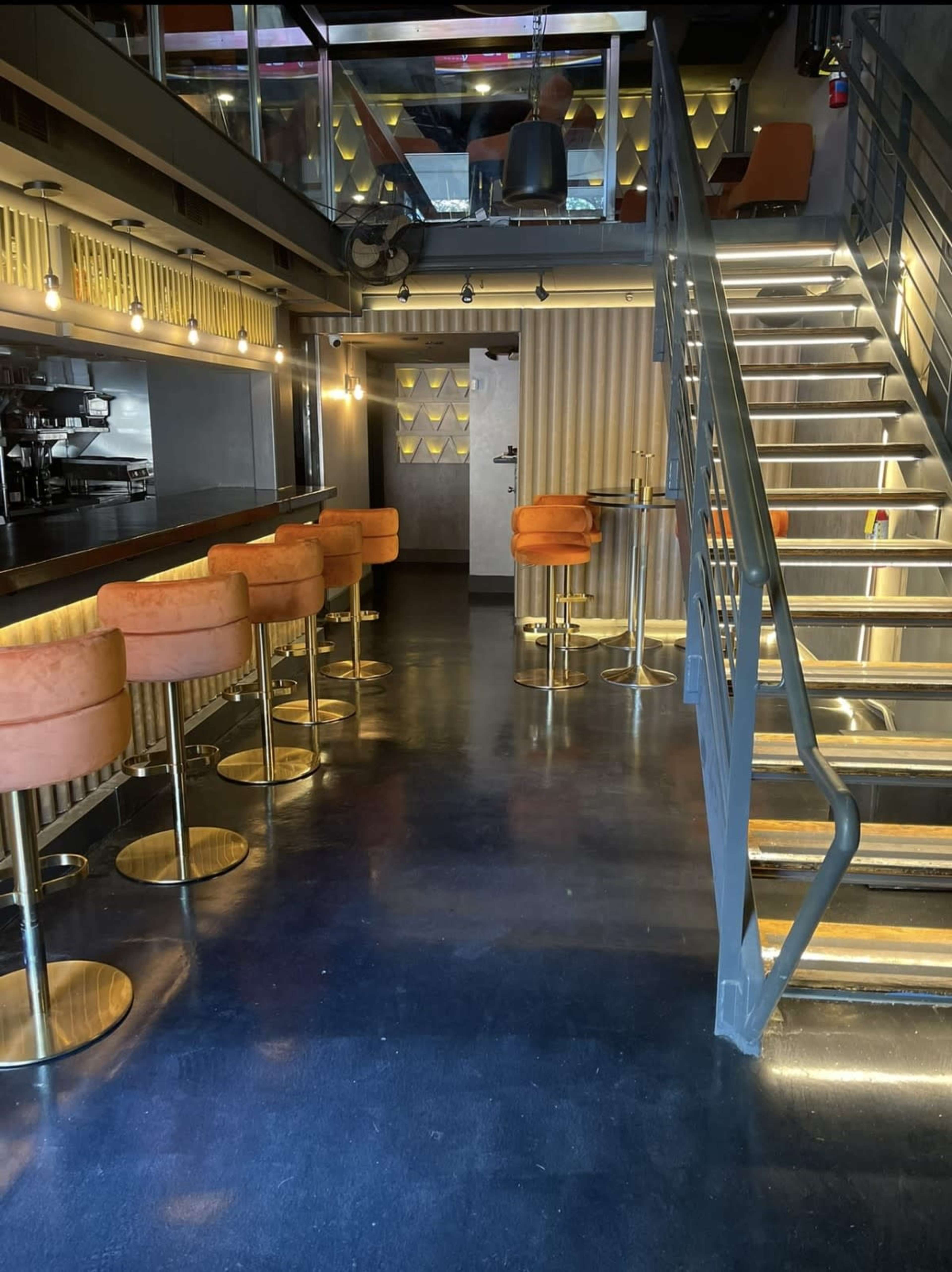 The image shows a modern interior of a restaurant with orange bar stools along a bar, a staircase leading to an upper level, and soft lighting.
