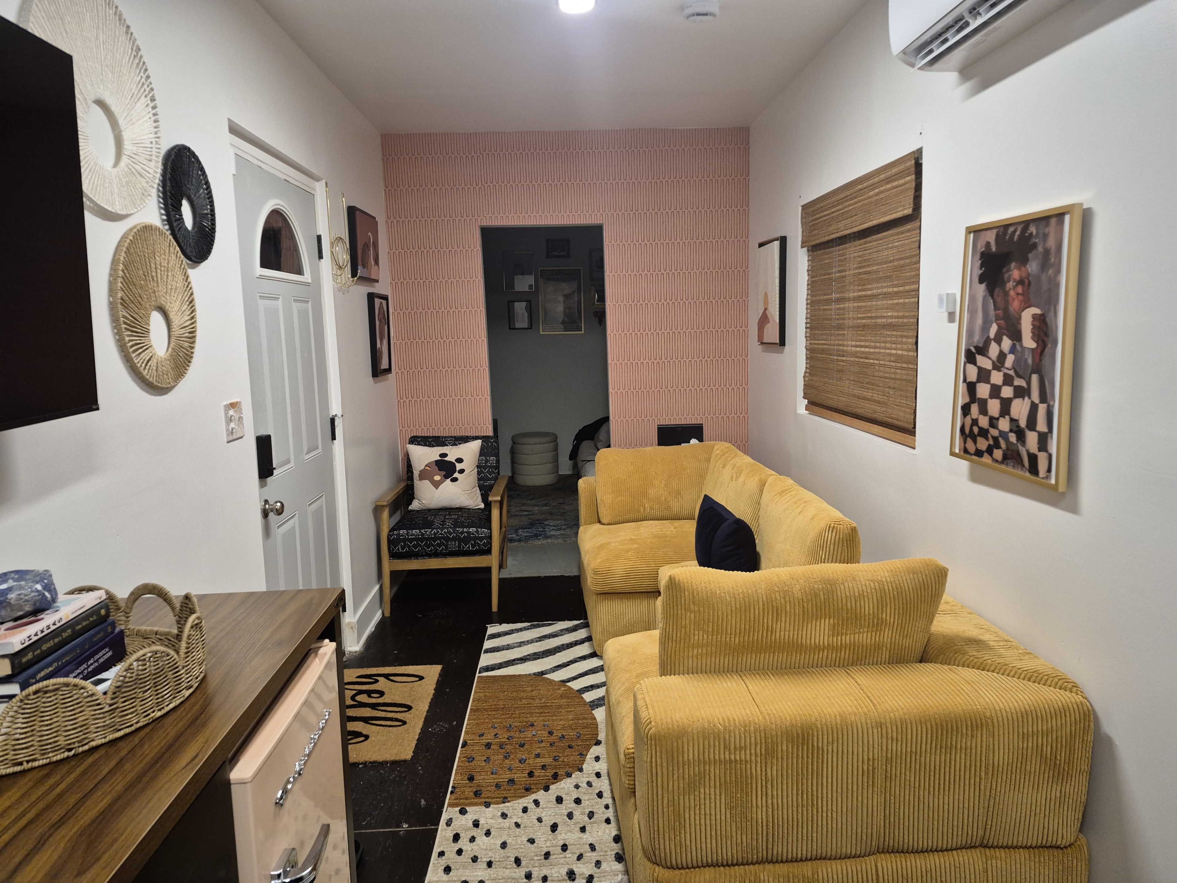 The image shows a small living room with yellow corduroy couches, a patterned rug, a TV on the wall, and framed artwork.