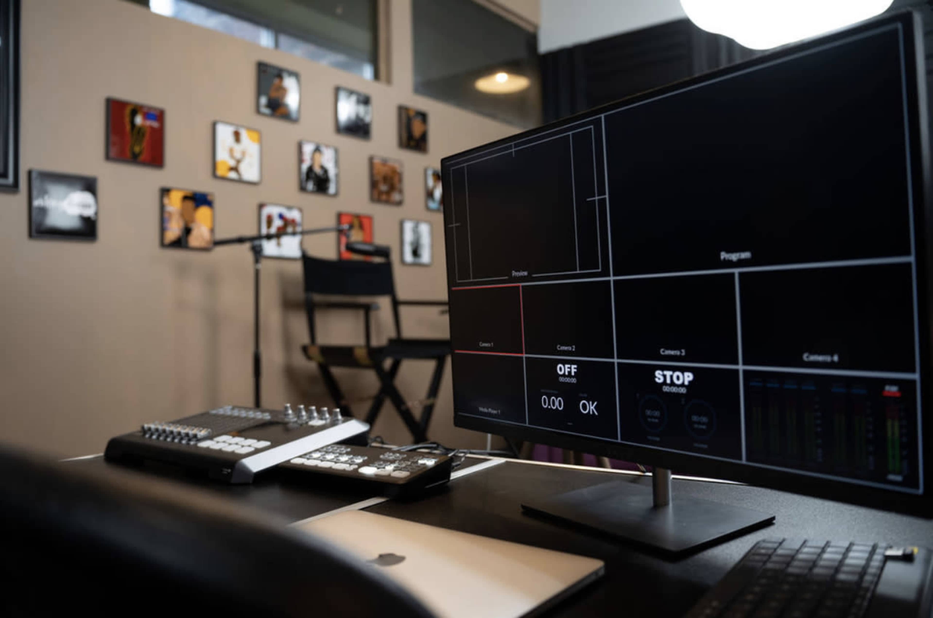 A computer monitor displaying video production controls is positioned on a desk in a studio adorned with framed artwork.