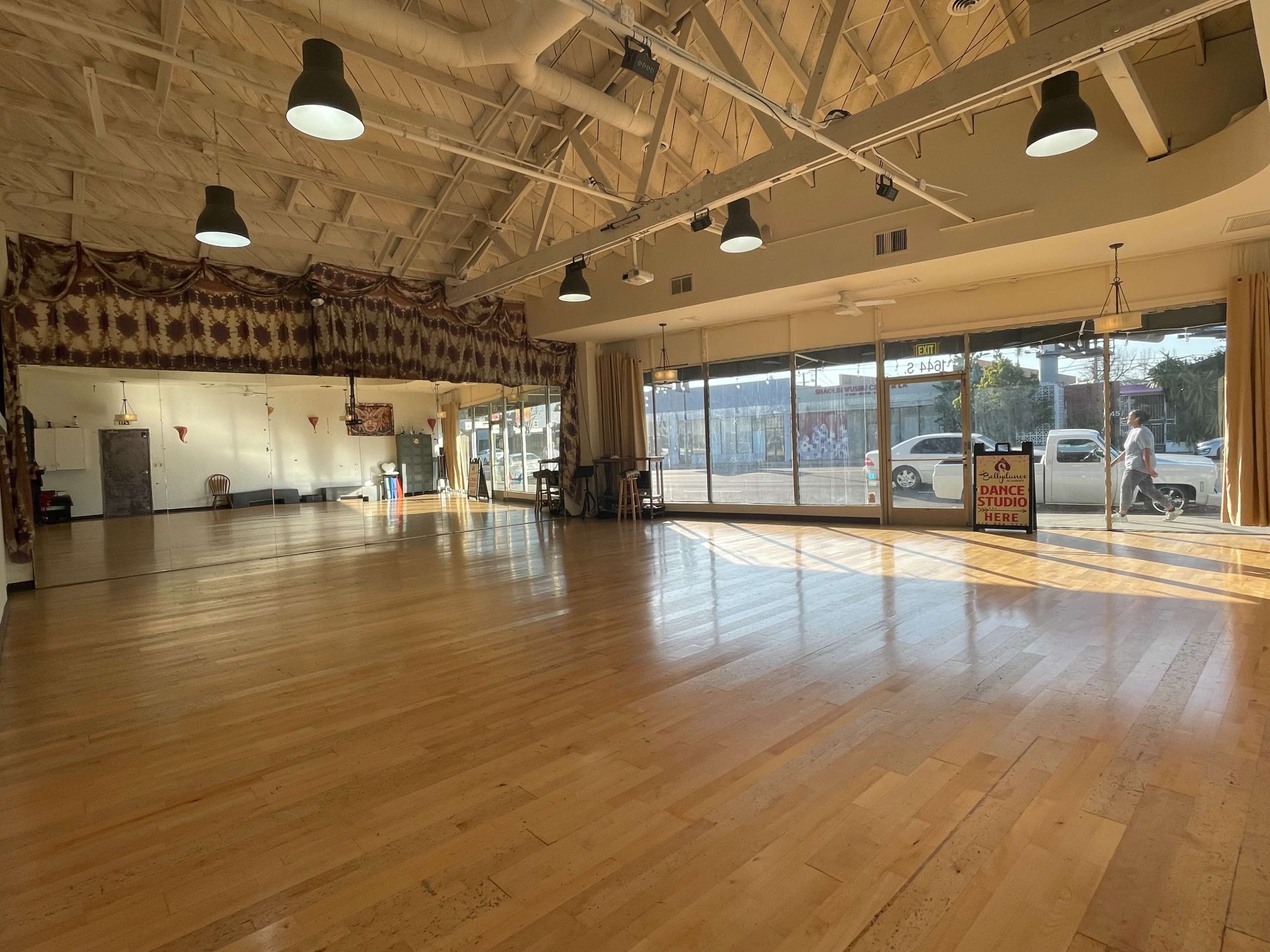 A brightly lit dance studio features large windows, polished wooden floors, and a wall draped with fabric.