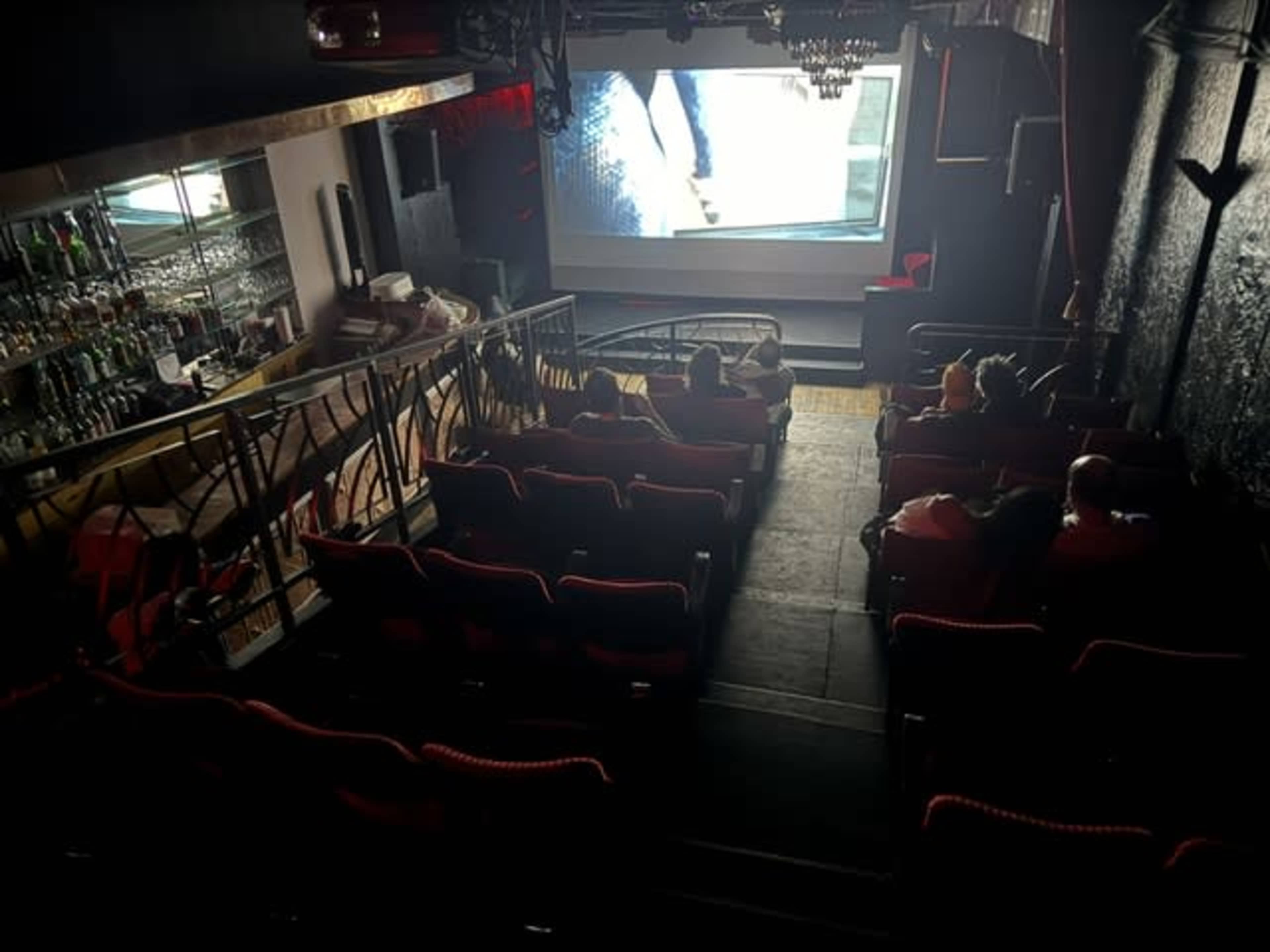 The image shows an empty small theater with red seats and a large screen displaying a scene, while a bar area is visible in the background.