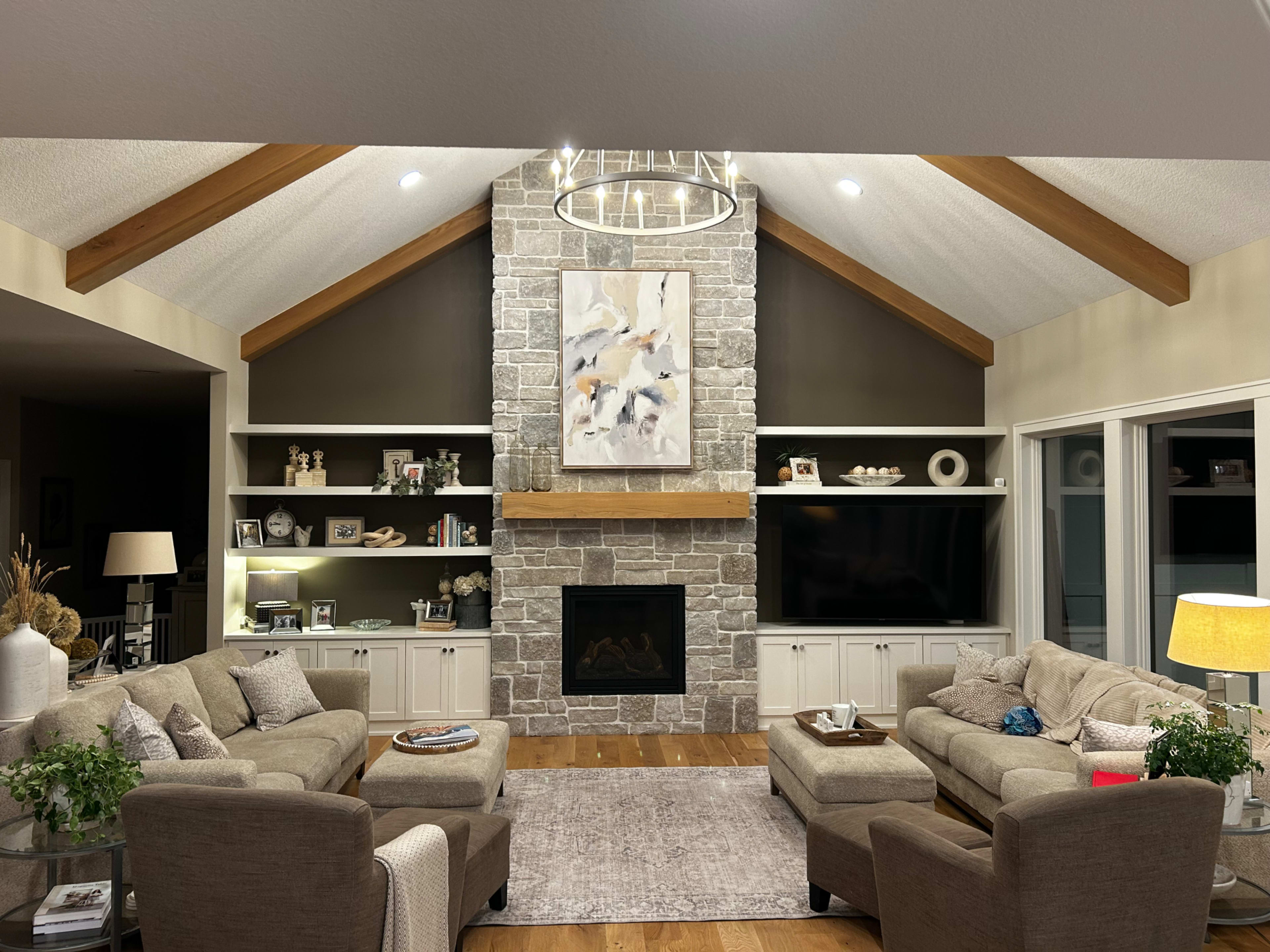 The image depicts a modern living room with a high, vaulted ceiling, featuring a stone fireplace and a large artwork above the mantel, surrounded by built-in shelving and comfortable seating.