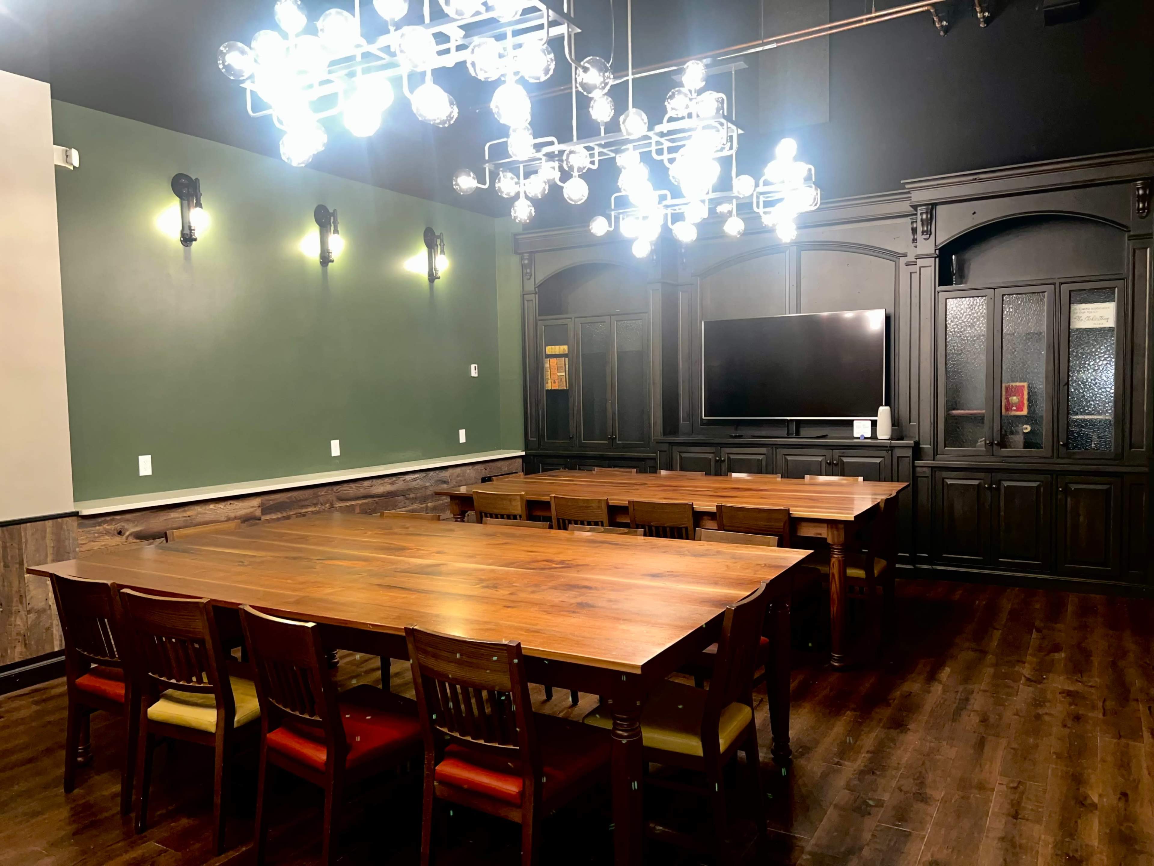The image shows a conference room with a long wooden table surrounded by chairs, a large television mounted on a dark cabinet, and a modern chandelier with glowing bulbs above.