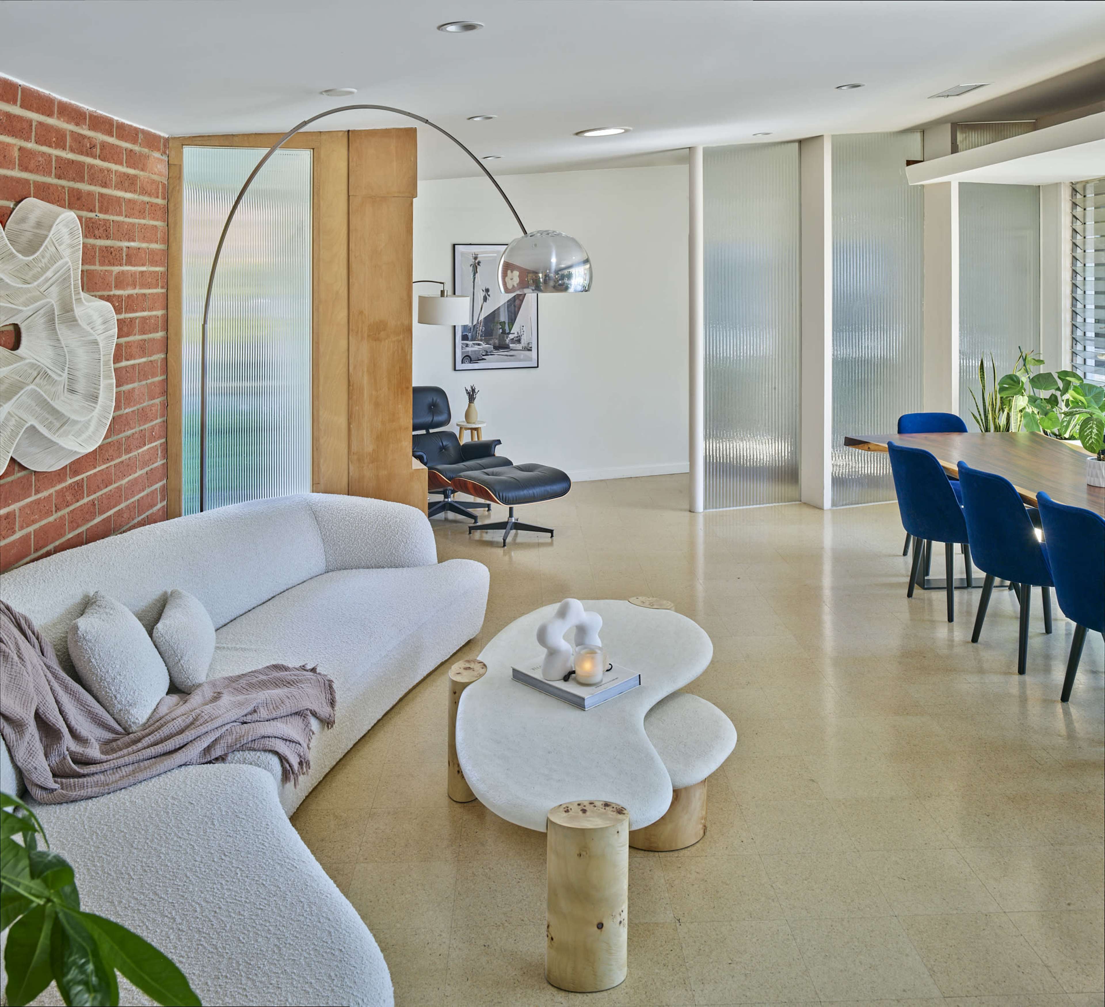 The scene shows a modern living space with a curved sofa, a light-colored coffee table made of stone, and a dining area featuring a wooden table and upholstered chairs.