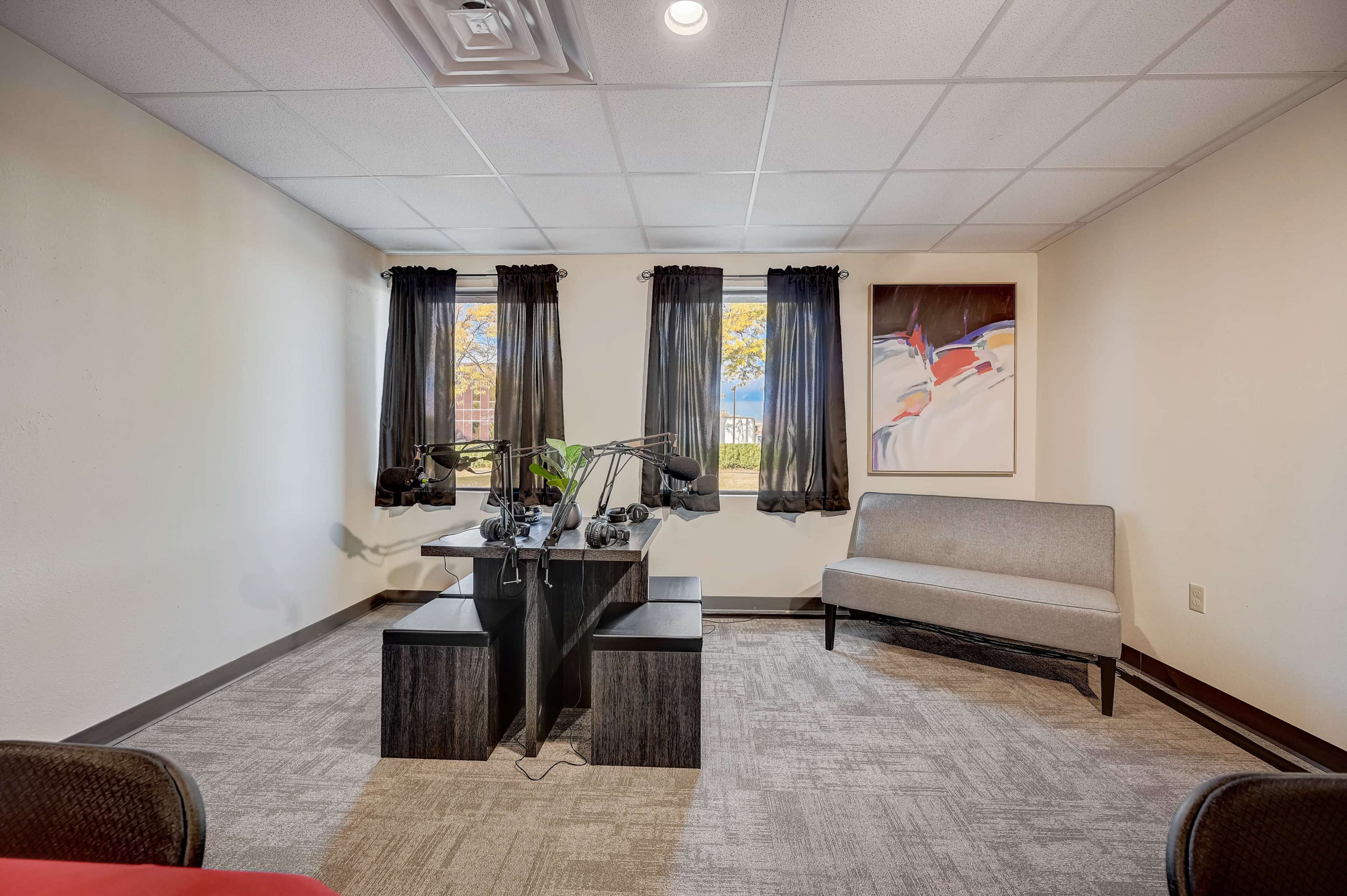 A small office space with a table and microphone setup, two windows with black curtains, and a gray sofa against the wall.