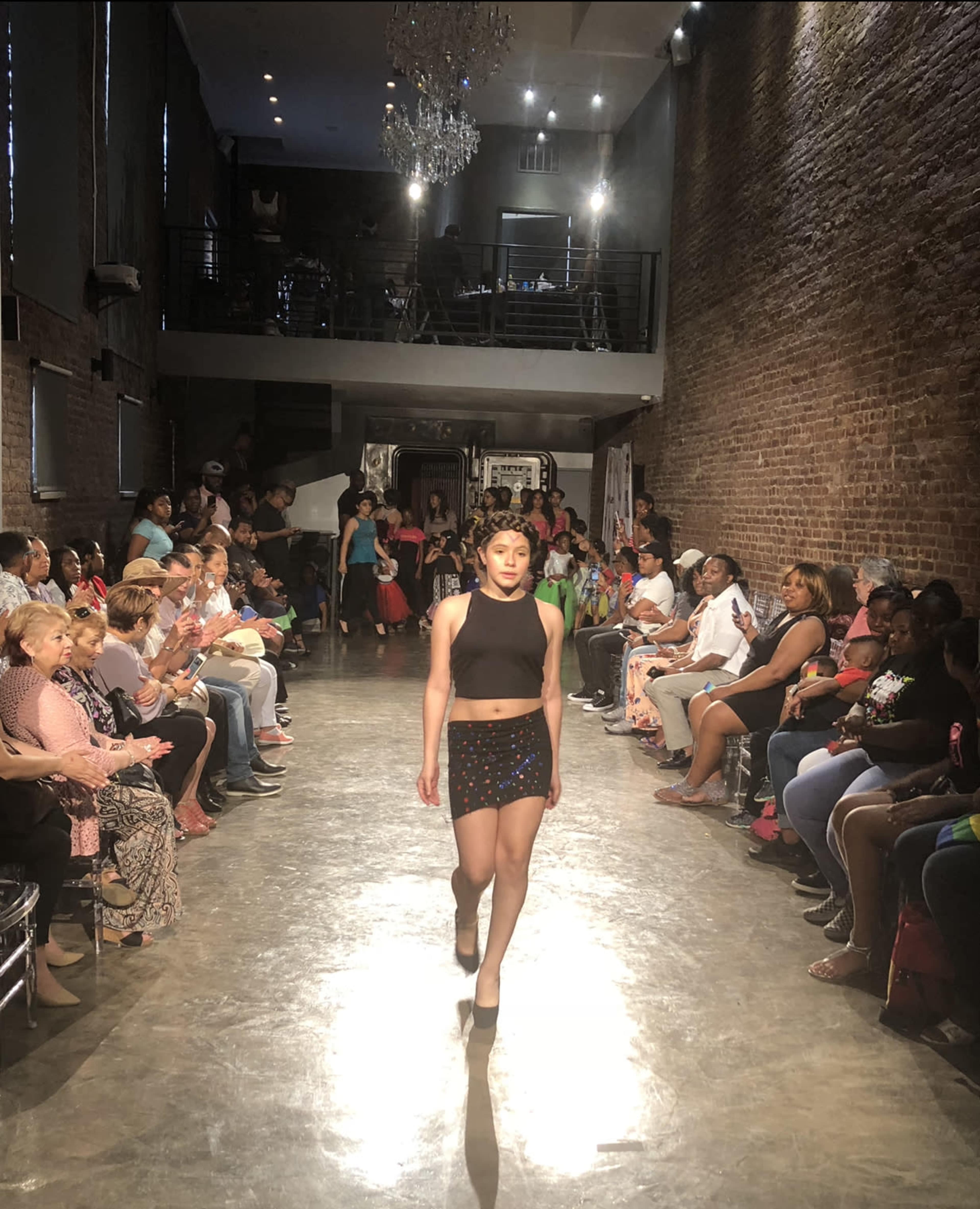 A model walks down a runway in front of a seated audience in a brick-walled venue.