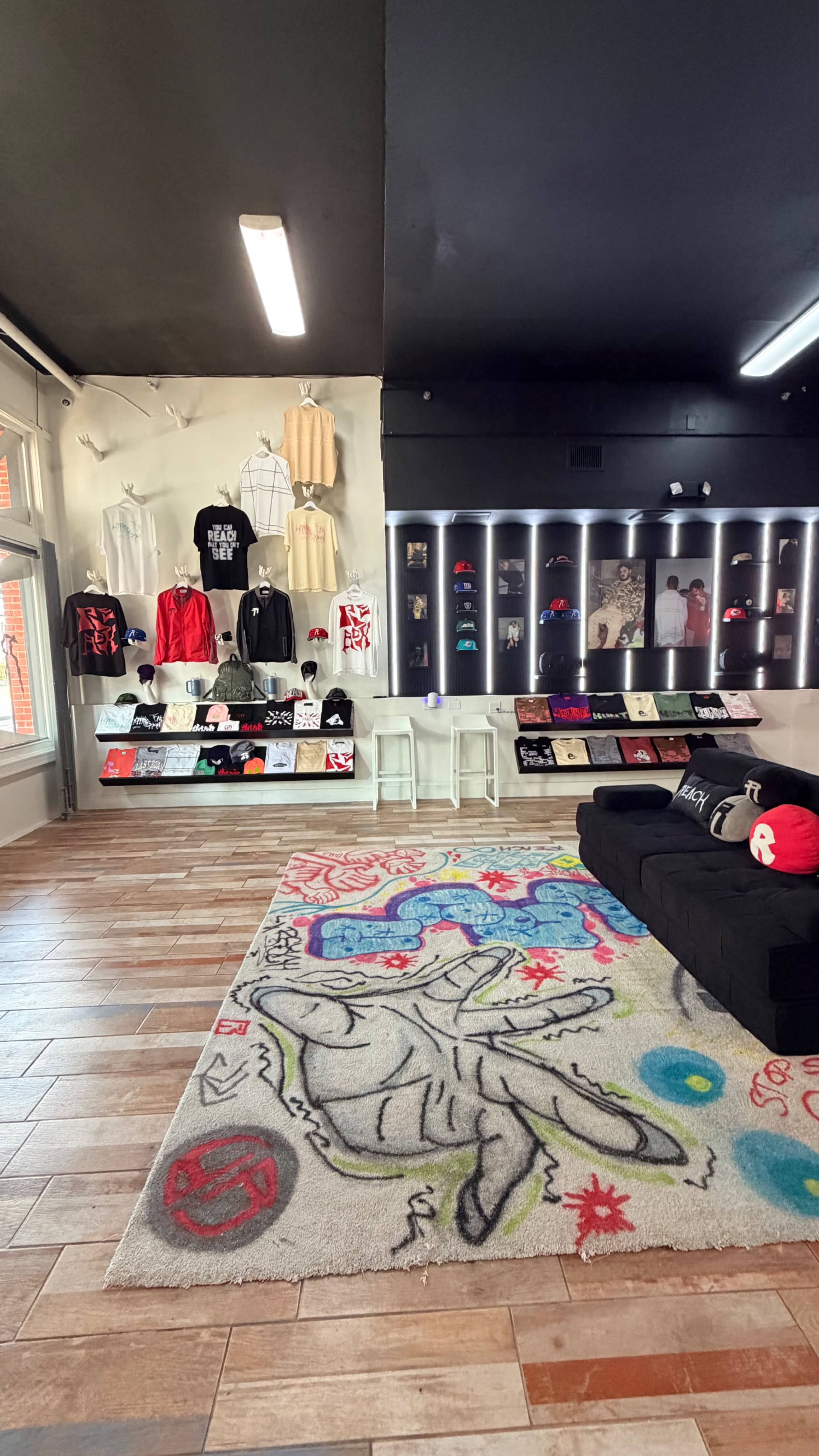 The image shows a retail space with a black couch, a colorful rug, and a display featuring various clothing items and accessories on the wall and shelves.