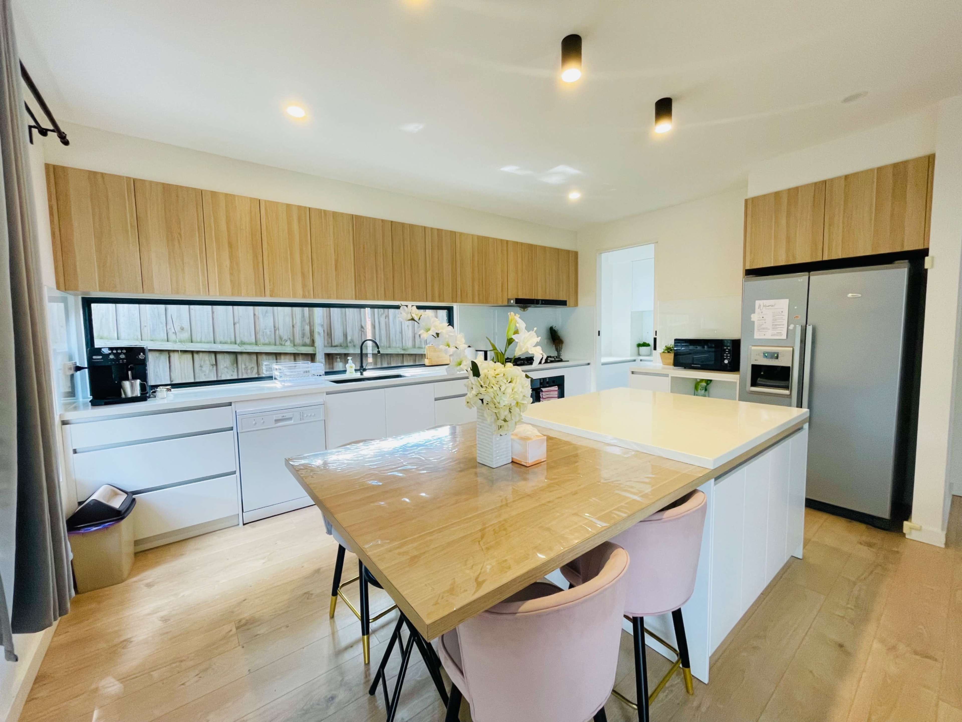 A modern kitchen features light wood cabinetry, white countertops, and a dining table with pink chairs, illuminated by overhead lighting.