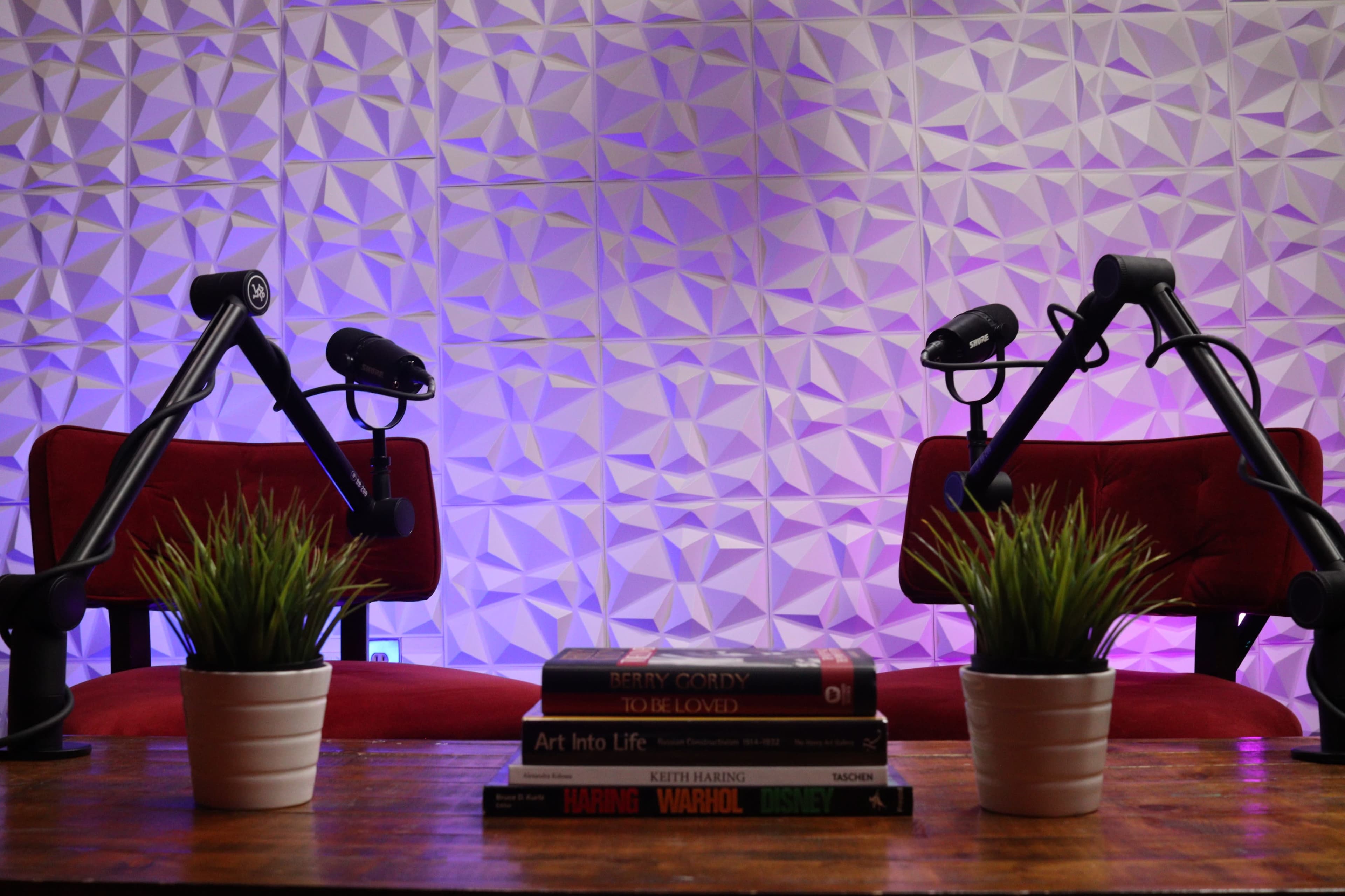 A small podcast or interview setup featuring two red chairs with microphone stands, a stack of books, and two potted plants on a wooden table against a textured, geometric wall backdrop.