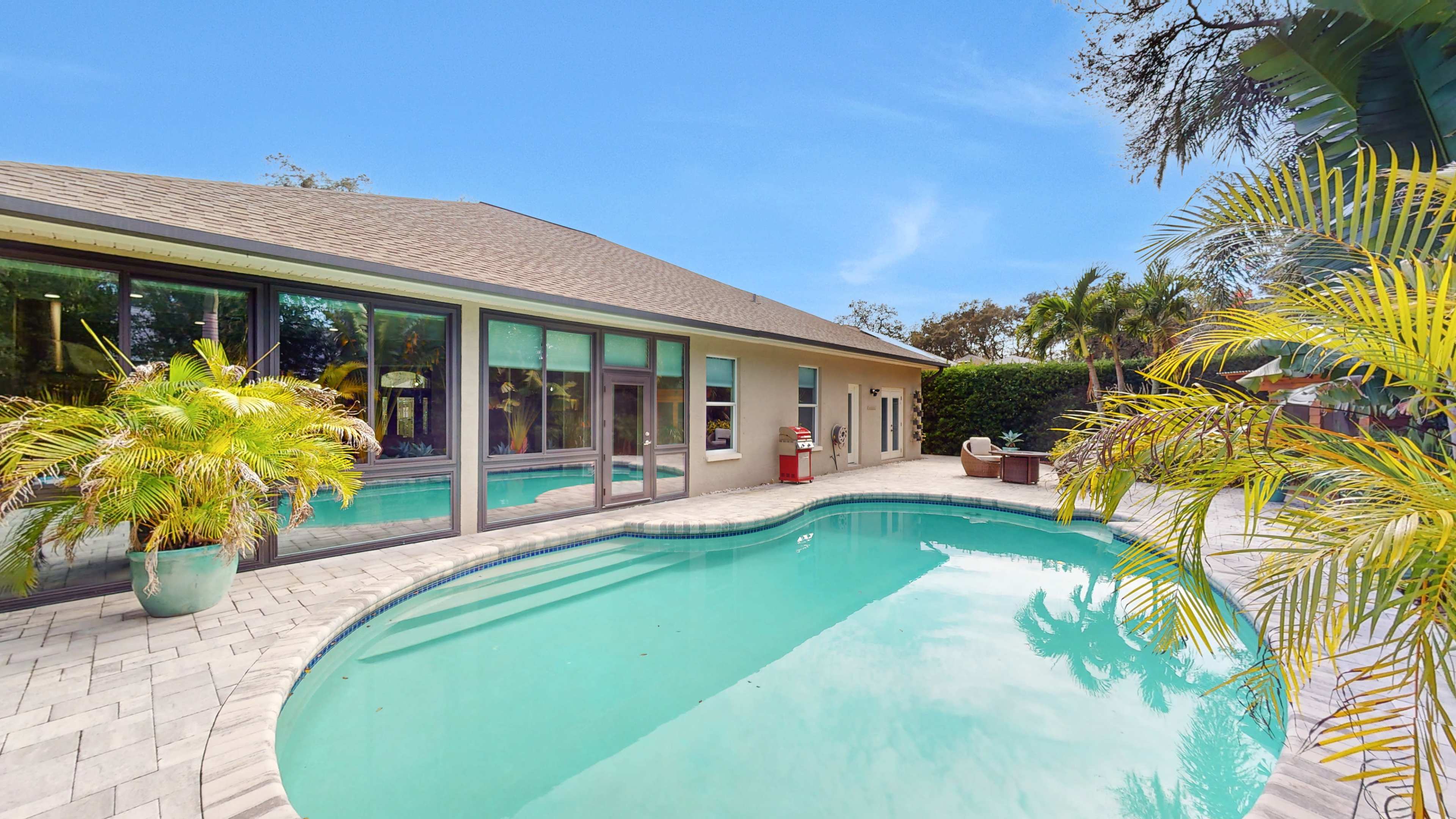 The image shows a well-maintained pool area surrounded by tropical plants and a modern home with large windows.