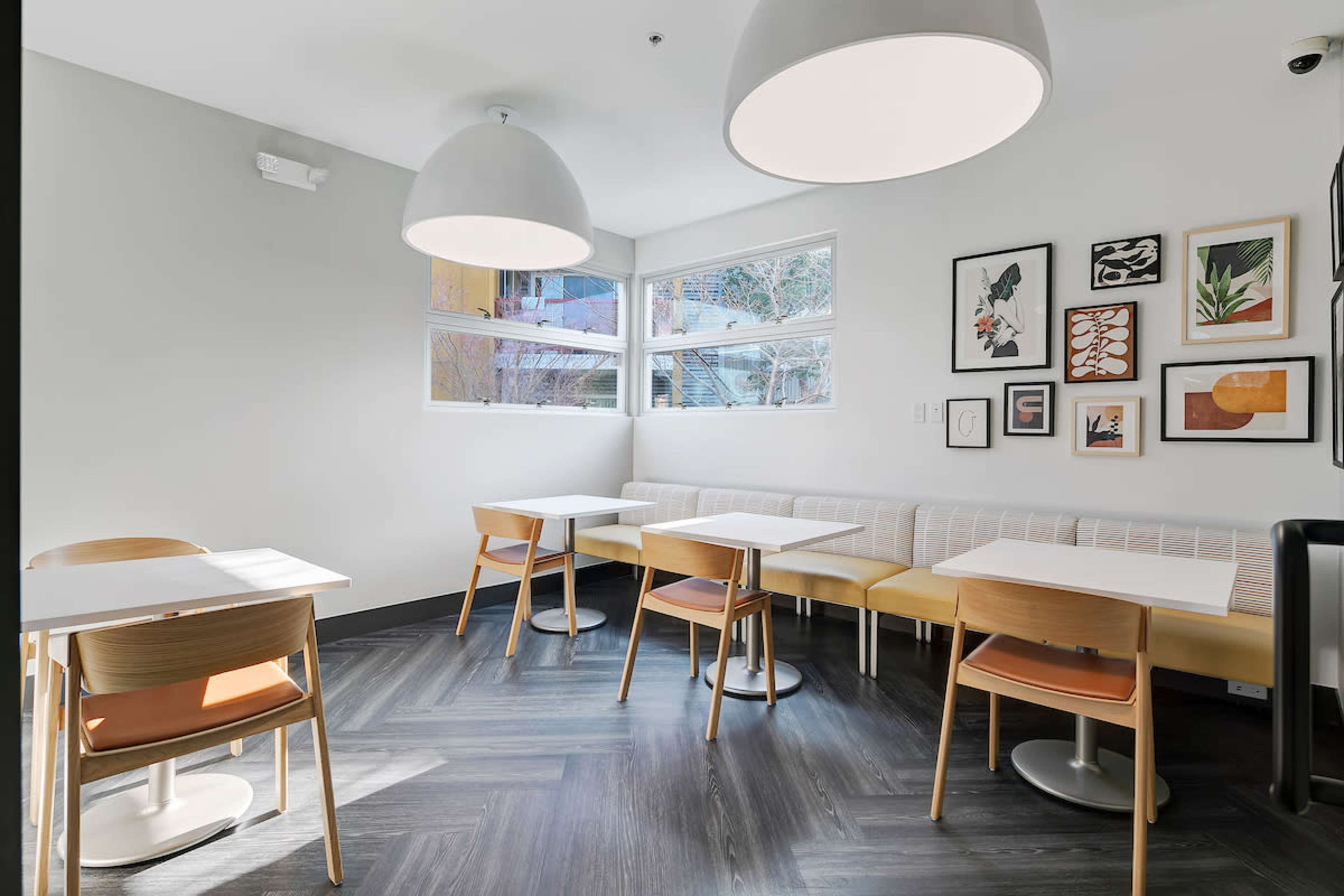 The image shows a bright, minimalist café interior featuring wooden tables, chairs, and large pendant lights, alongside a wall adorned with framed artwork.