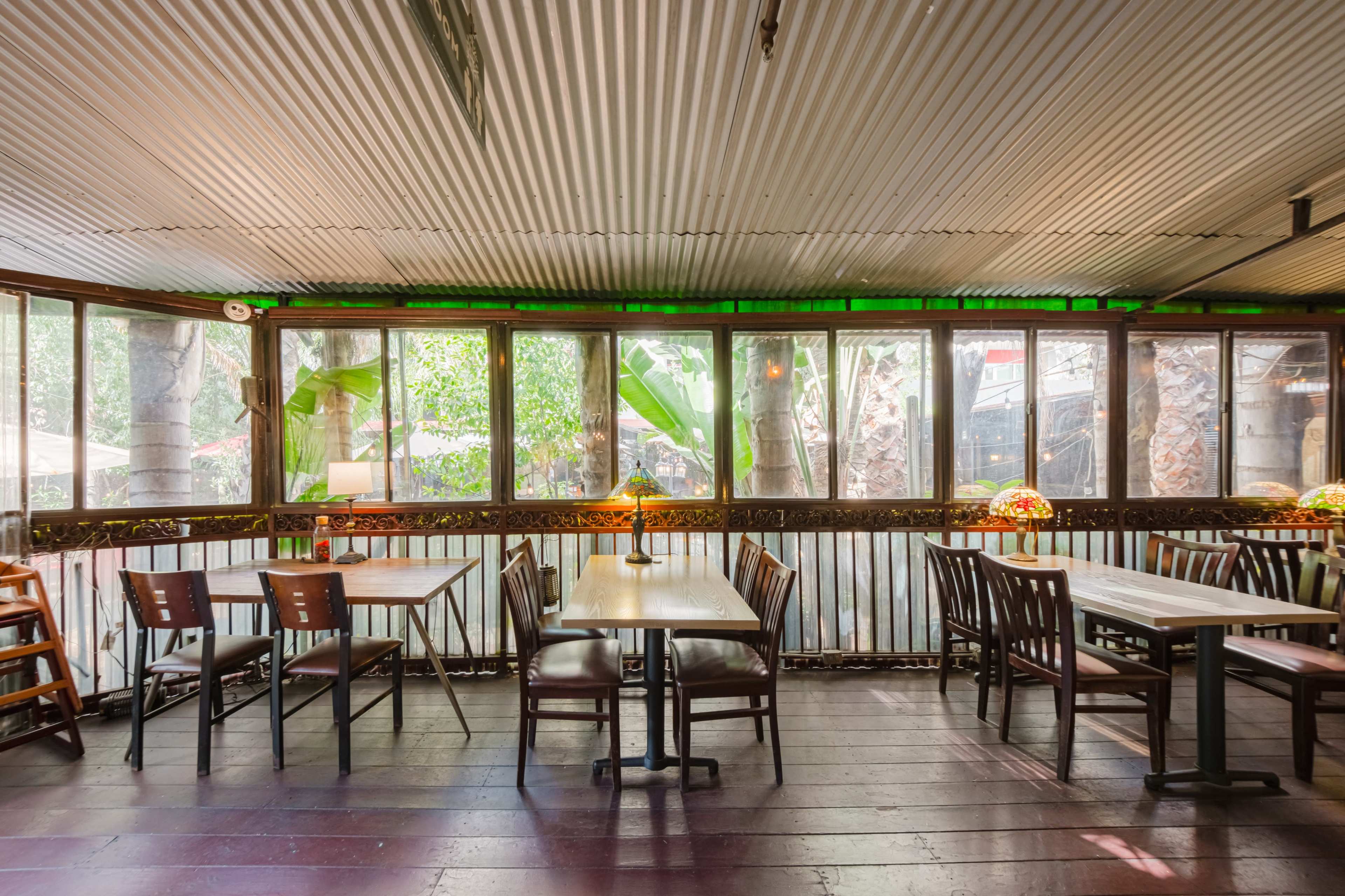 An empty restaurant interior with wooden tables and chairs situated by large windows that overlook greenery.