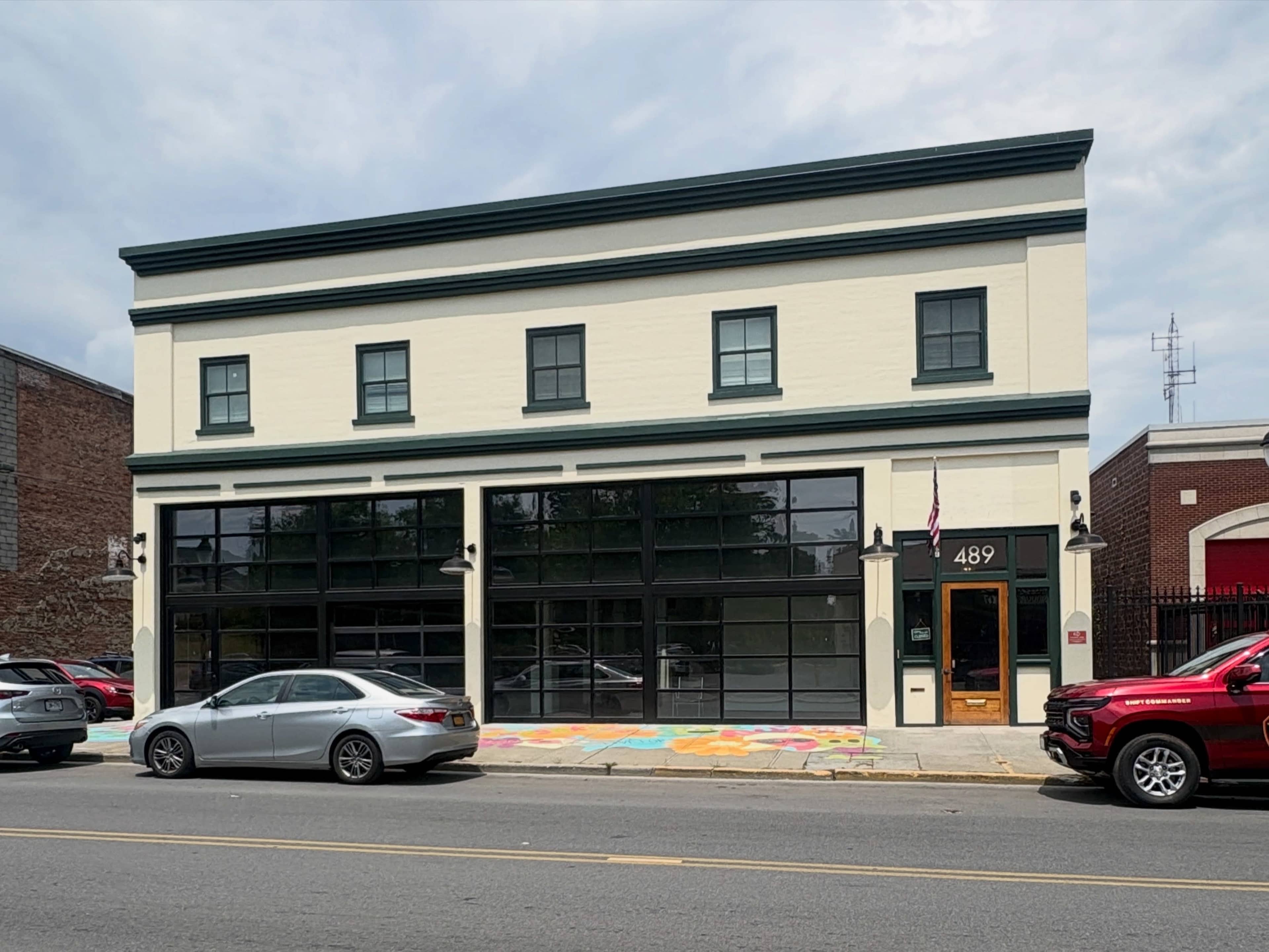 The image shows a two-story building with large windows and an American flag, located on a street with parked cars in front.