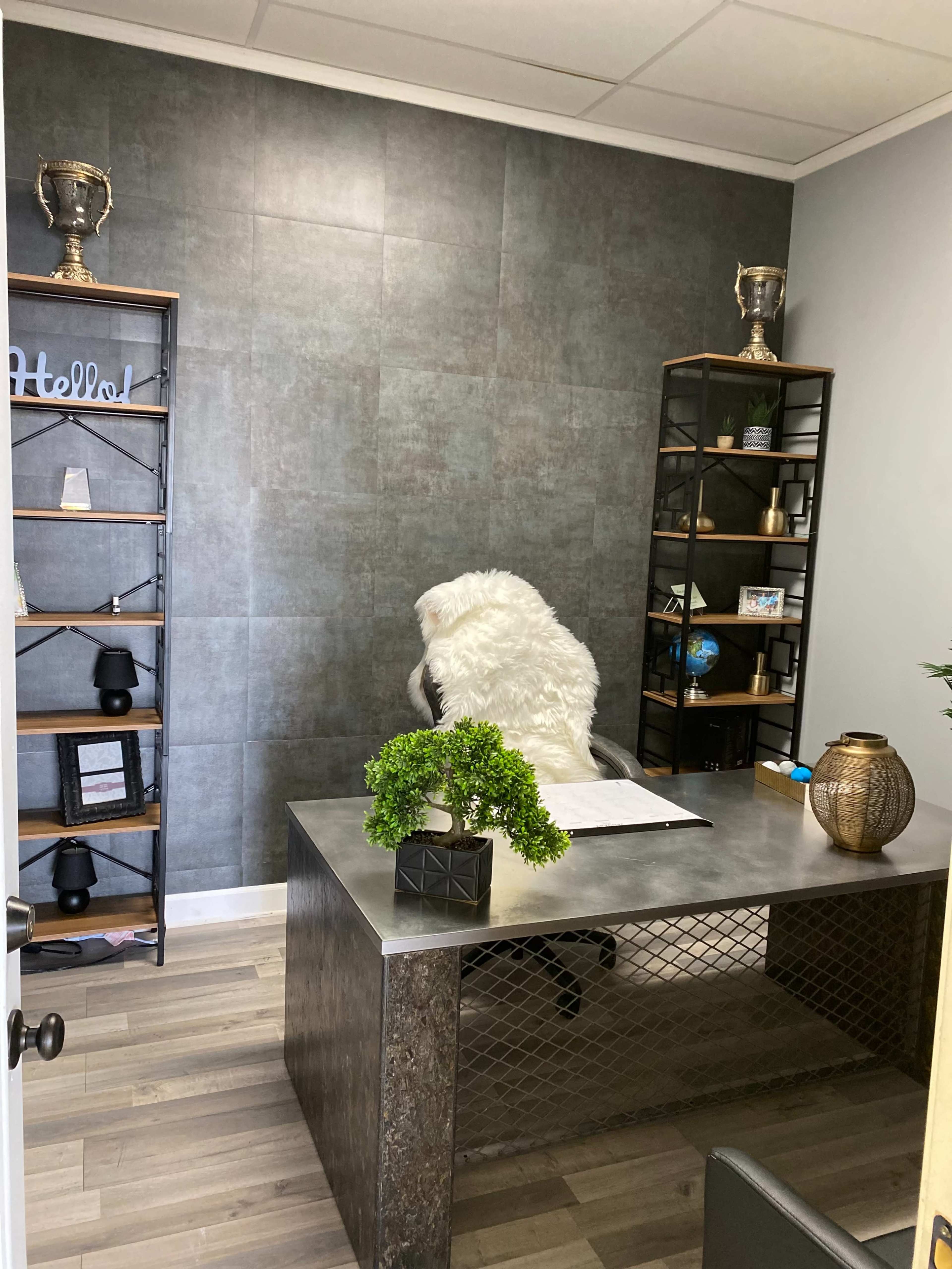 The image shows a modern office space with a metal desk, a chair covered in a white fur throw, and black shelving units displaying decorative items.