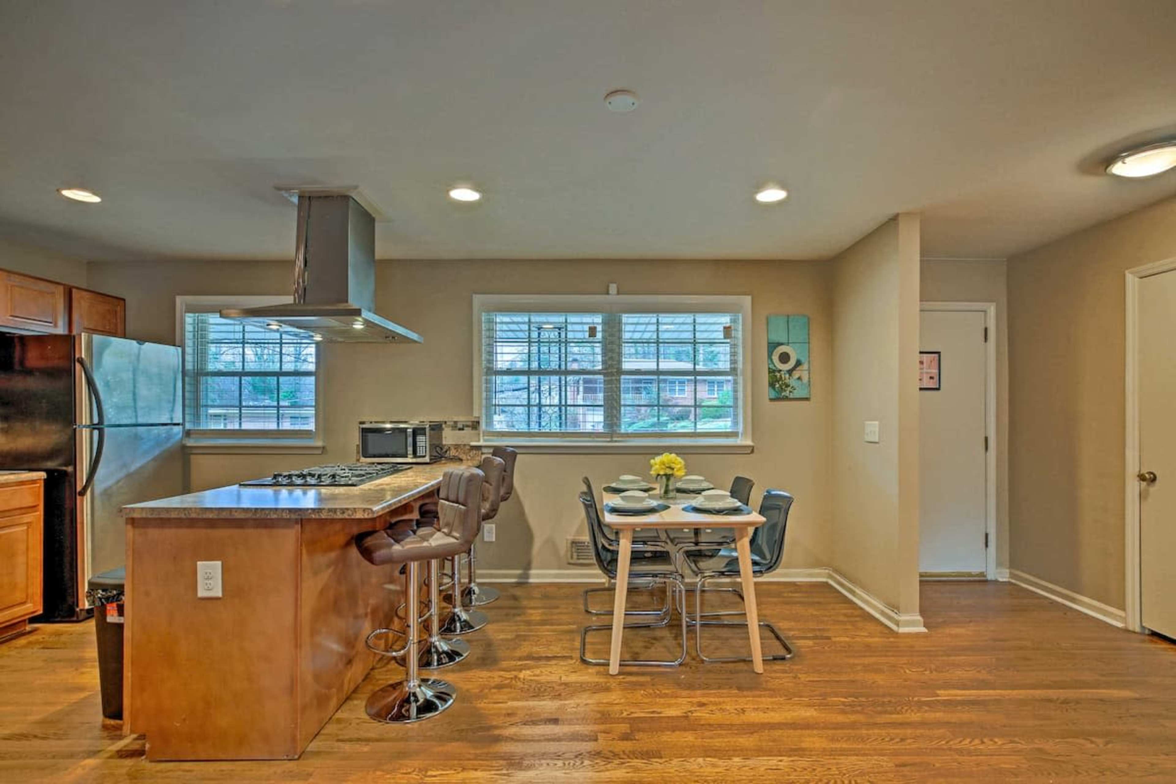 A kitchen with a central island, bar stools, a dining table set for four, and windows allowing natural light.