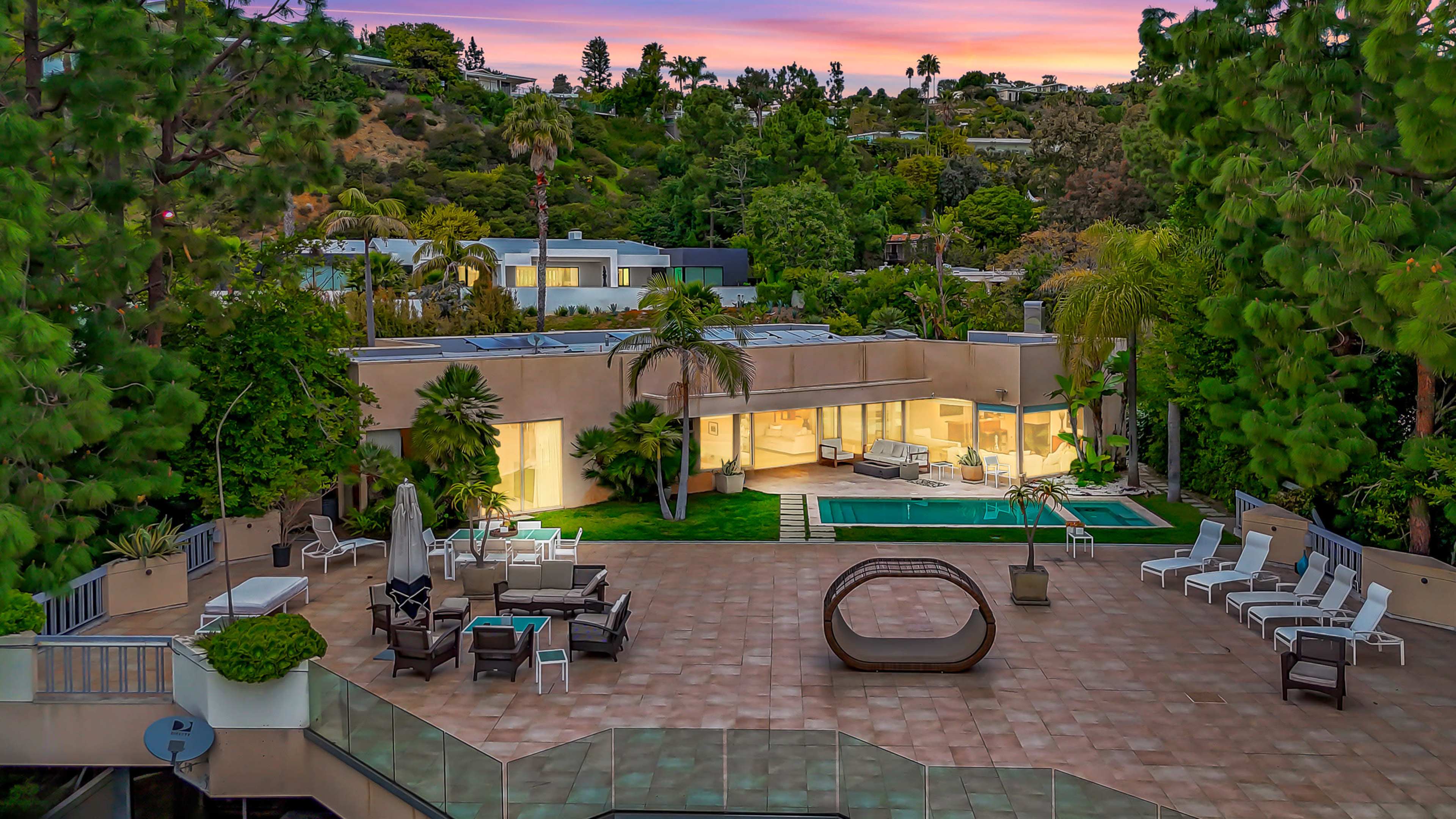 A modern house with large glass windows, surrounded by trees and outdoor furniture, alongside a swimming pool and a patio area.