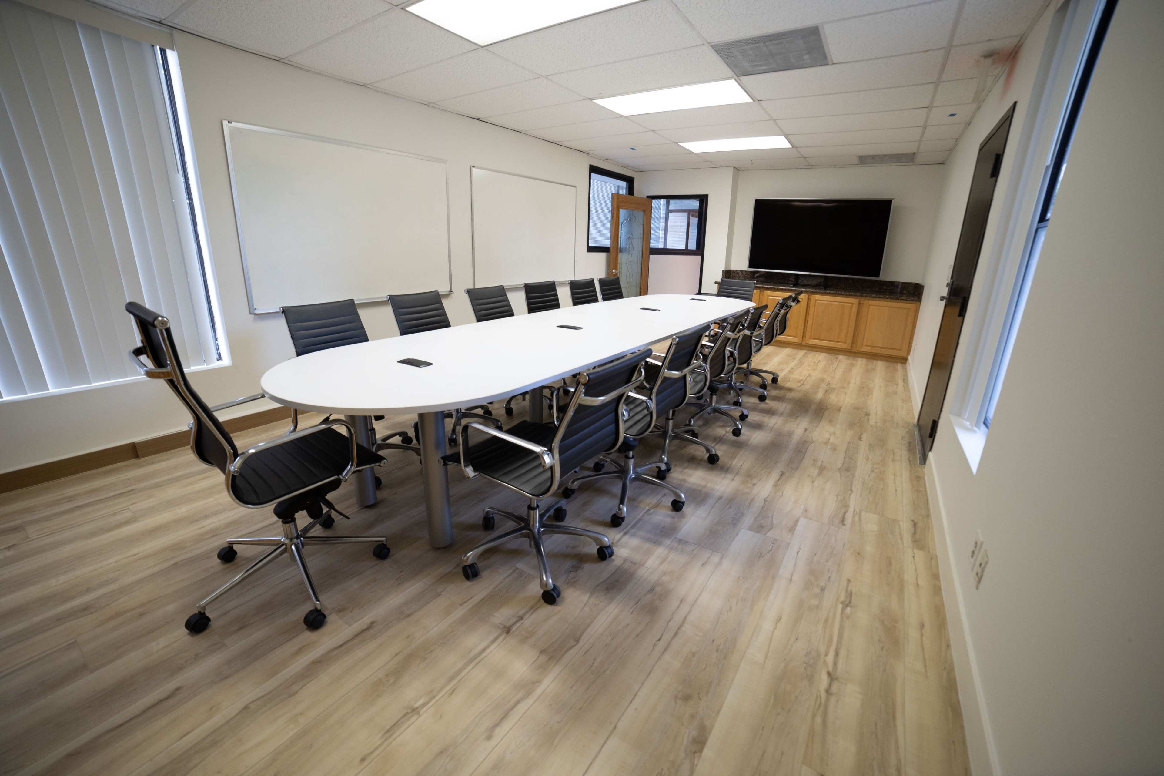 A large conference room features a long white table surrounded by black office chairs, with a television and whiteboards along the walls.