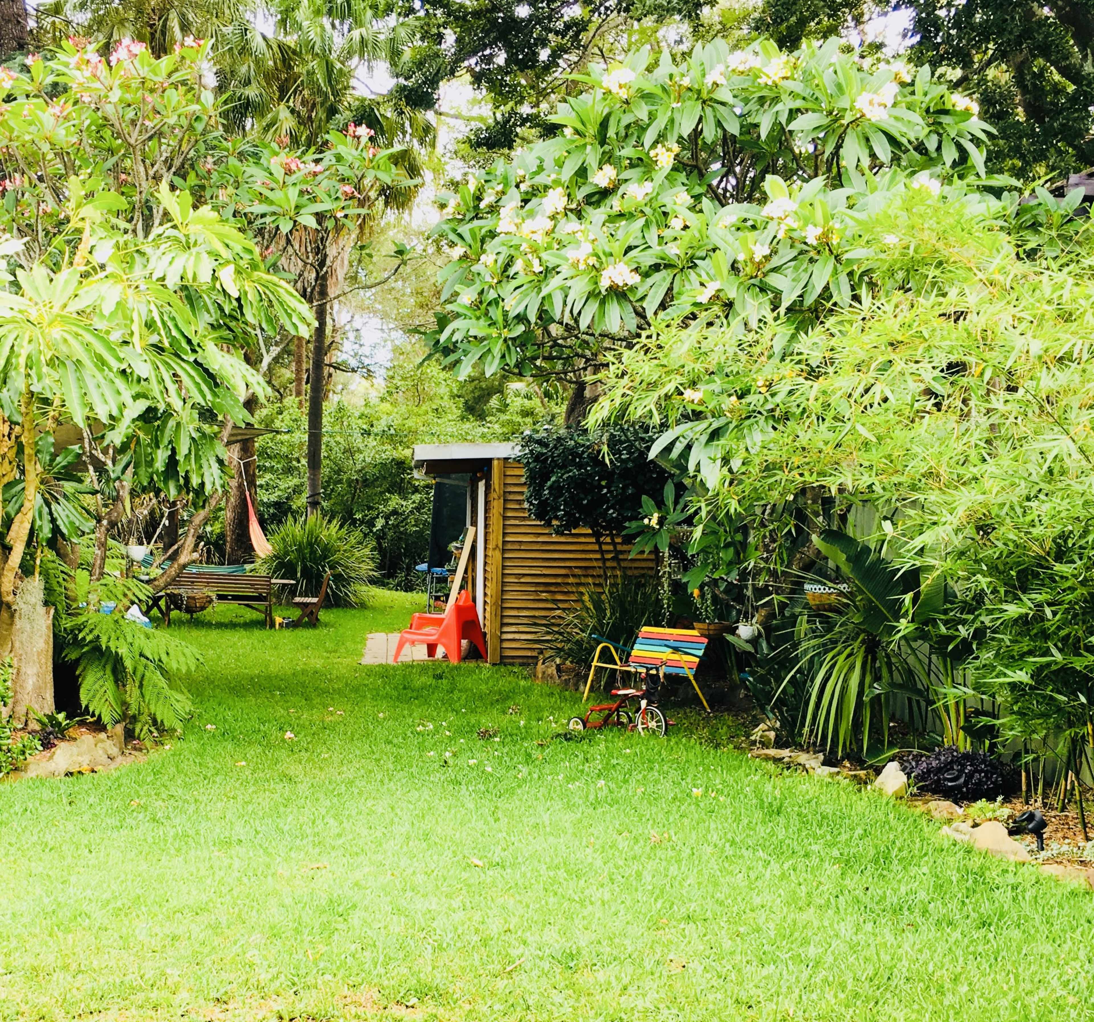A lush garden features green grass, tropical plants, and a small shed, with a children's toy and red plastic chair visible along the pathway.