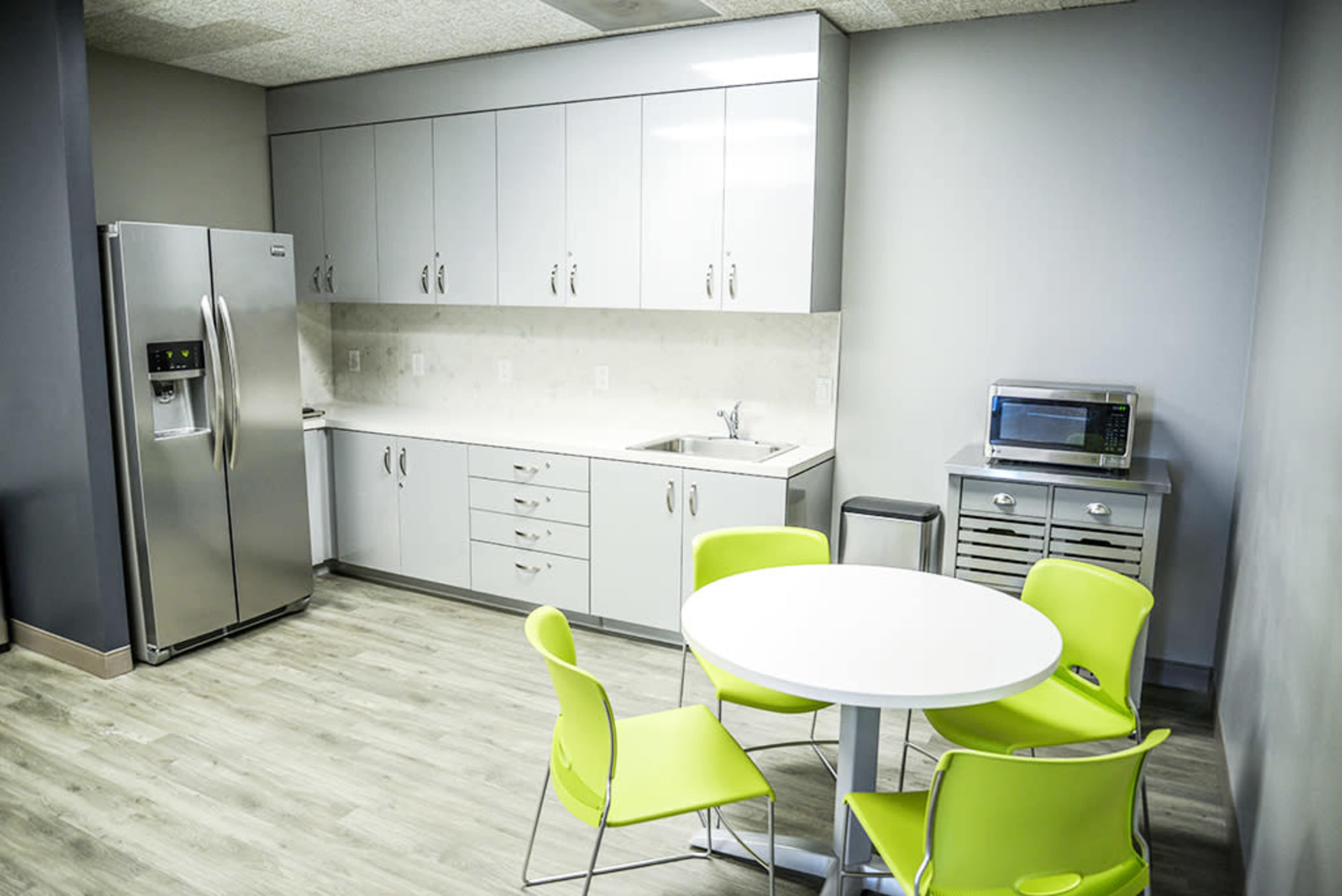 A modern kitchenette with stainless steel appliances, a white countertop, and a round table surrounded by bright green chairs.