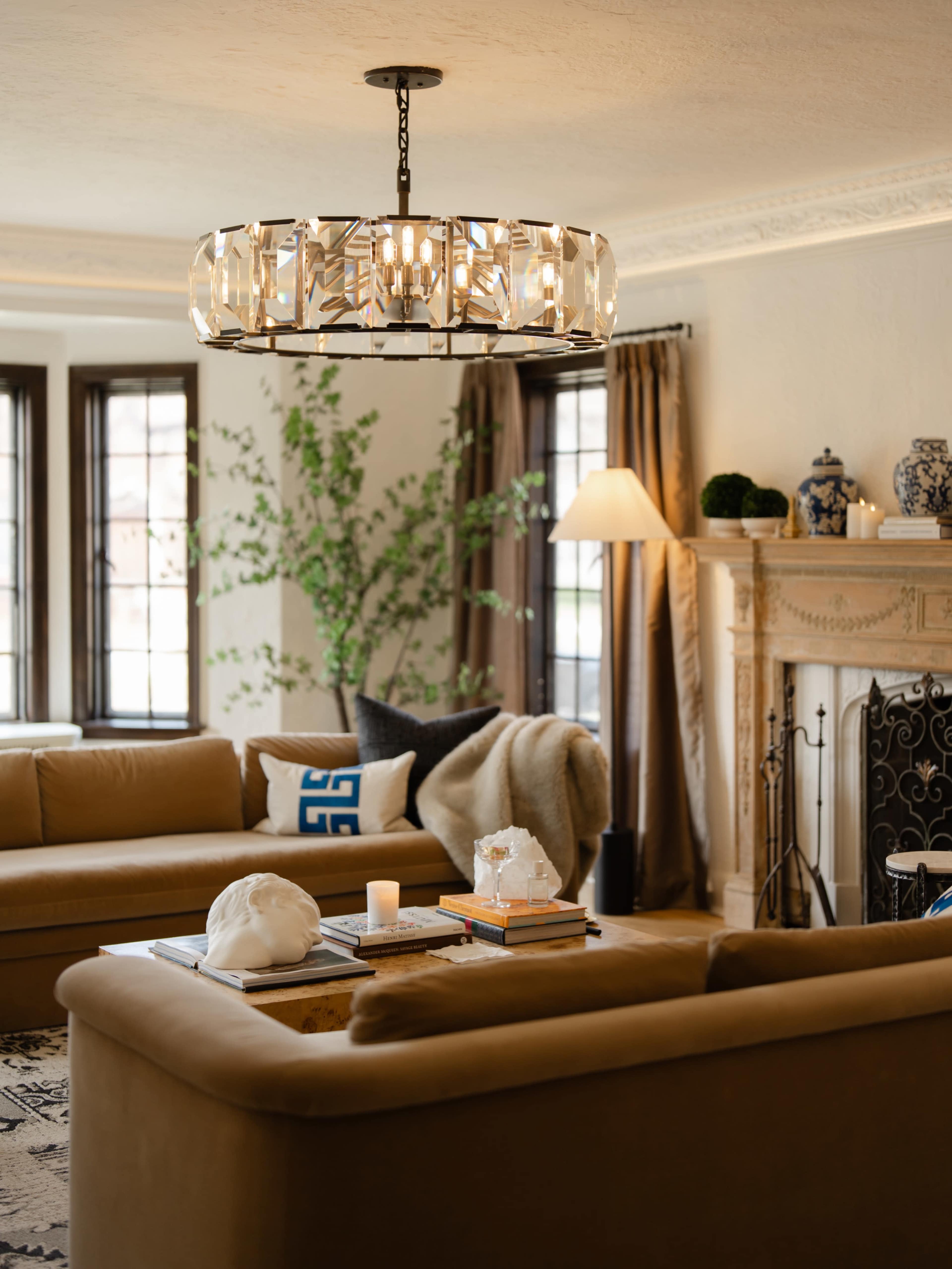 The image shows a spacious living room with a modern chandelier, plush beige sofas, a decorative fireplace, and a tall plant beside a floor lamp.