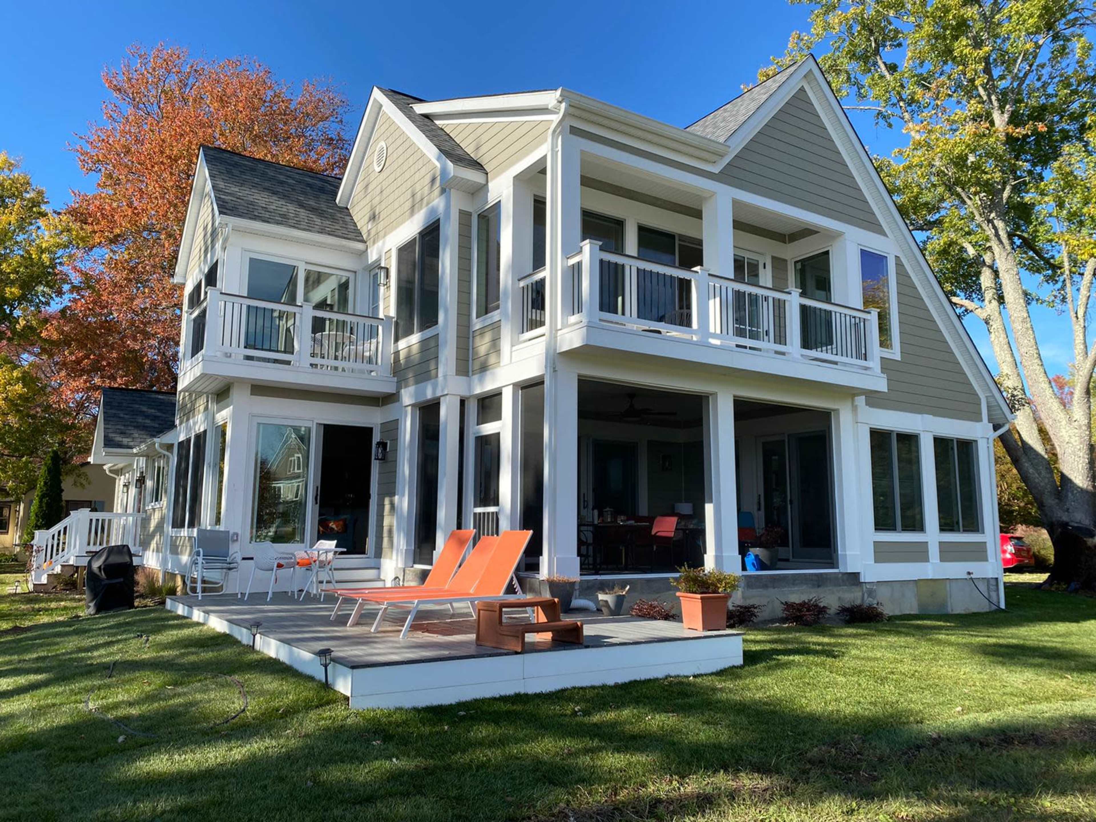 A modern two-story house with large windows, a deck, and orange lounge chairs in a grassy area, framed by trees displaying fall colors.