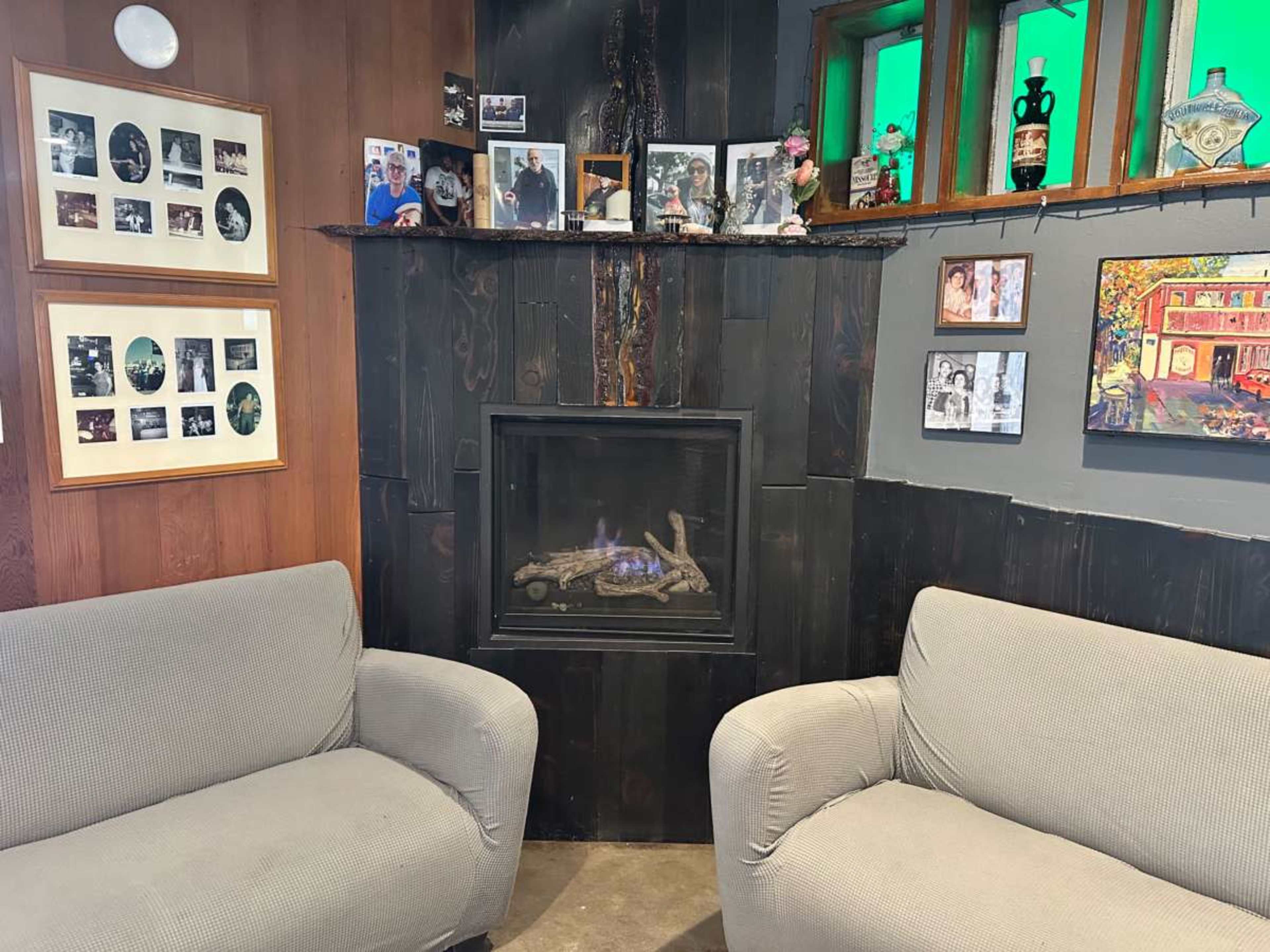 The image shows a cozy corner of a room with two beige armchairs facing a modern electric fireplace, surrounded by framed photographs and decorations on the walls.