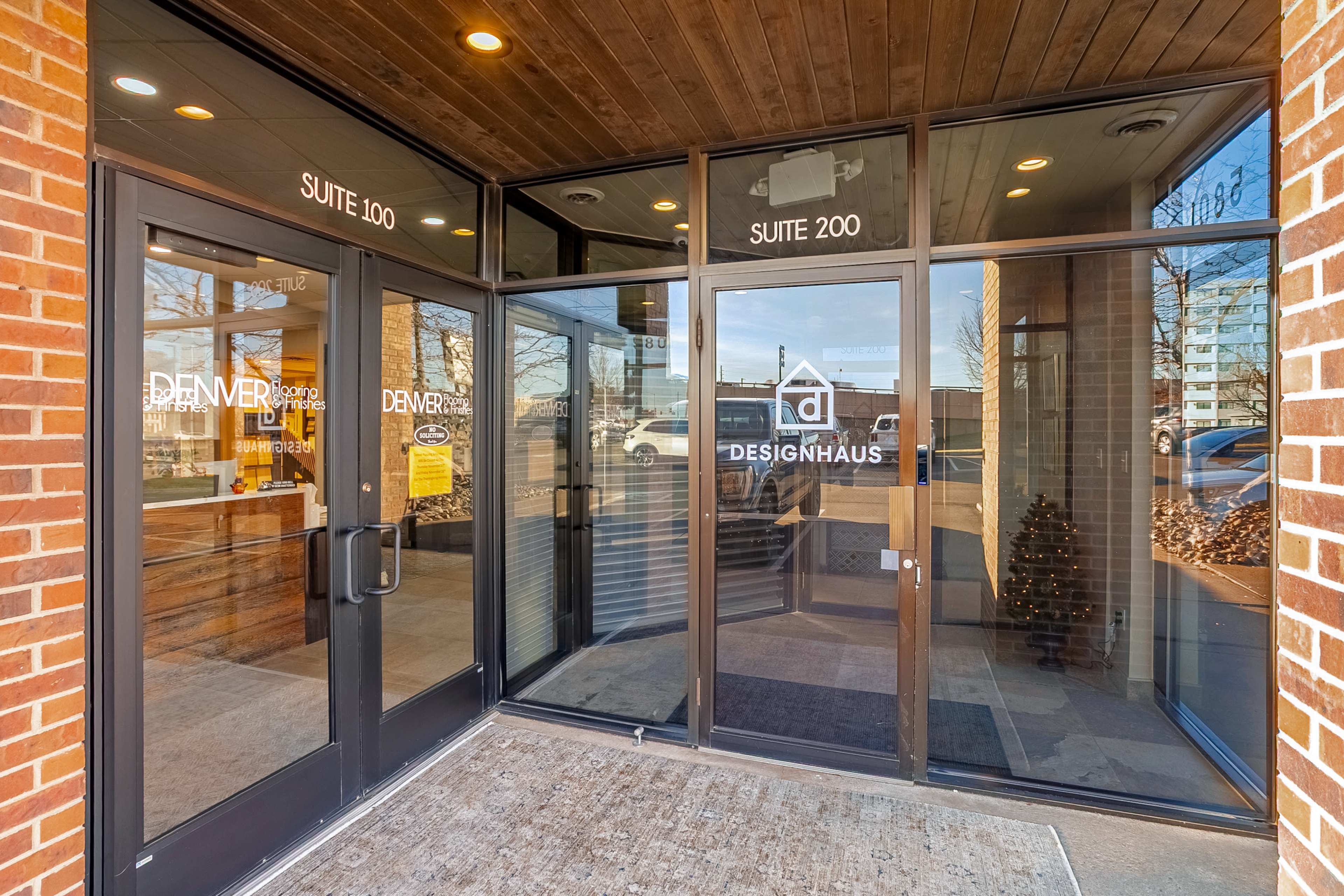 The image shows the entrance of a building featuring glass doors with signage indicating "DENVER" and "DESIGNHAUS" alongside suite numbers.