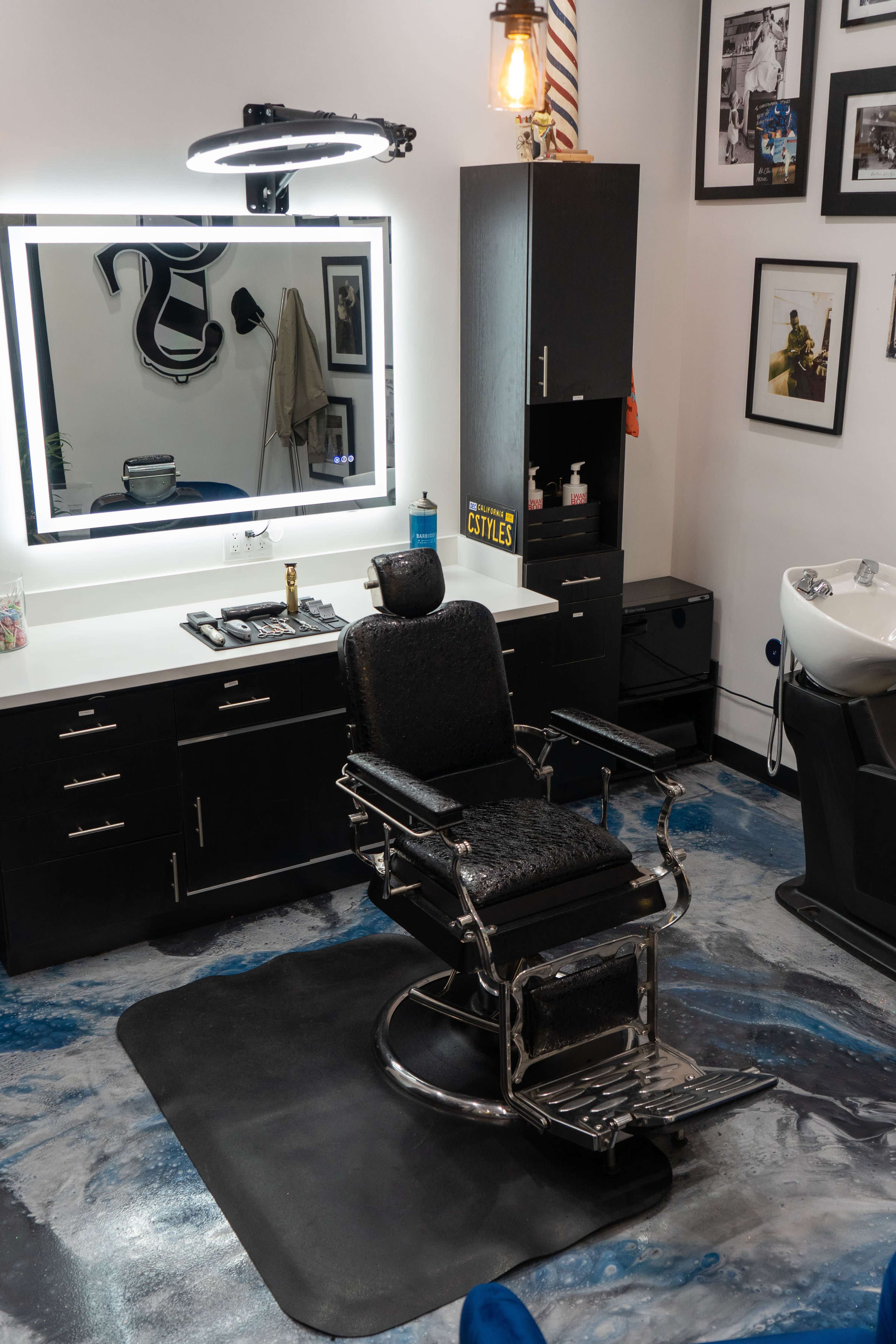 The interior of a barbershop features a black barber chair, a large illuminated mirror, and various grooming products on shelves.
