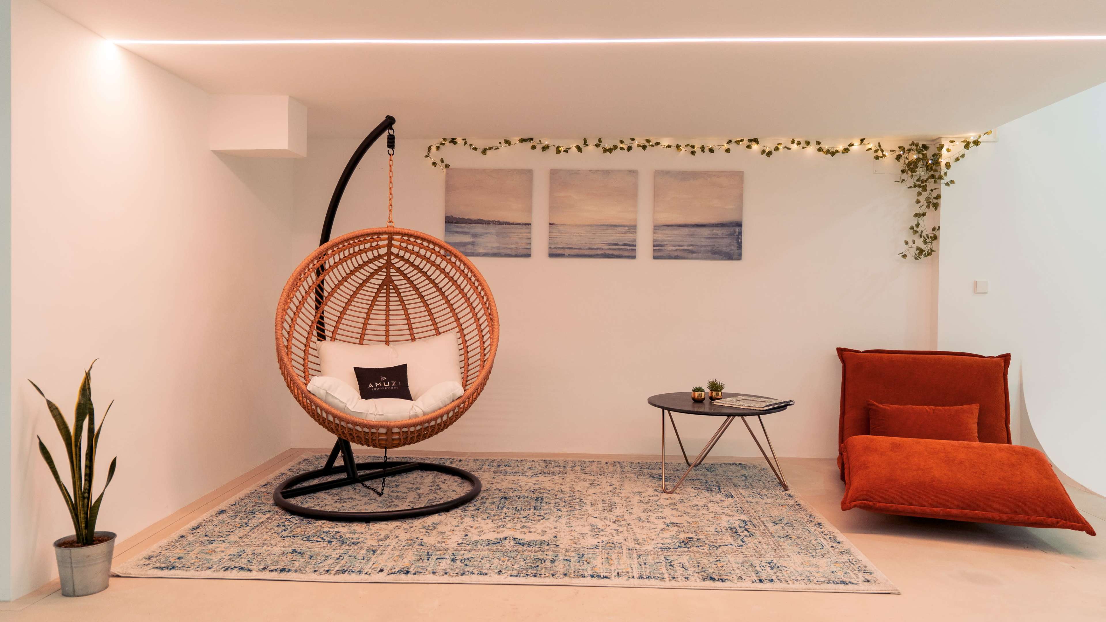 The image features a modern interior with a hanging rattan chair, a cozy orange lounge chair, a round coffee table, and decorative wall art.