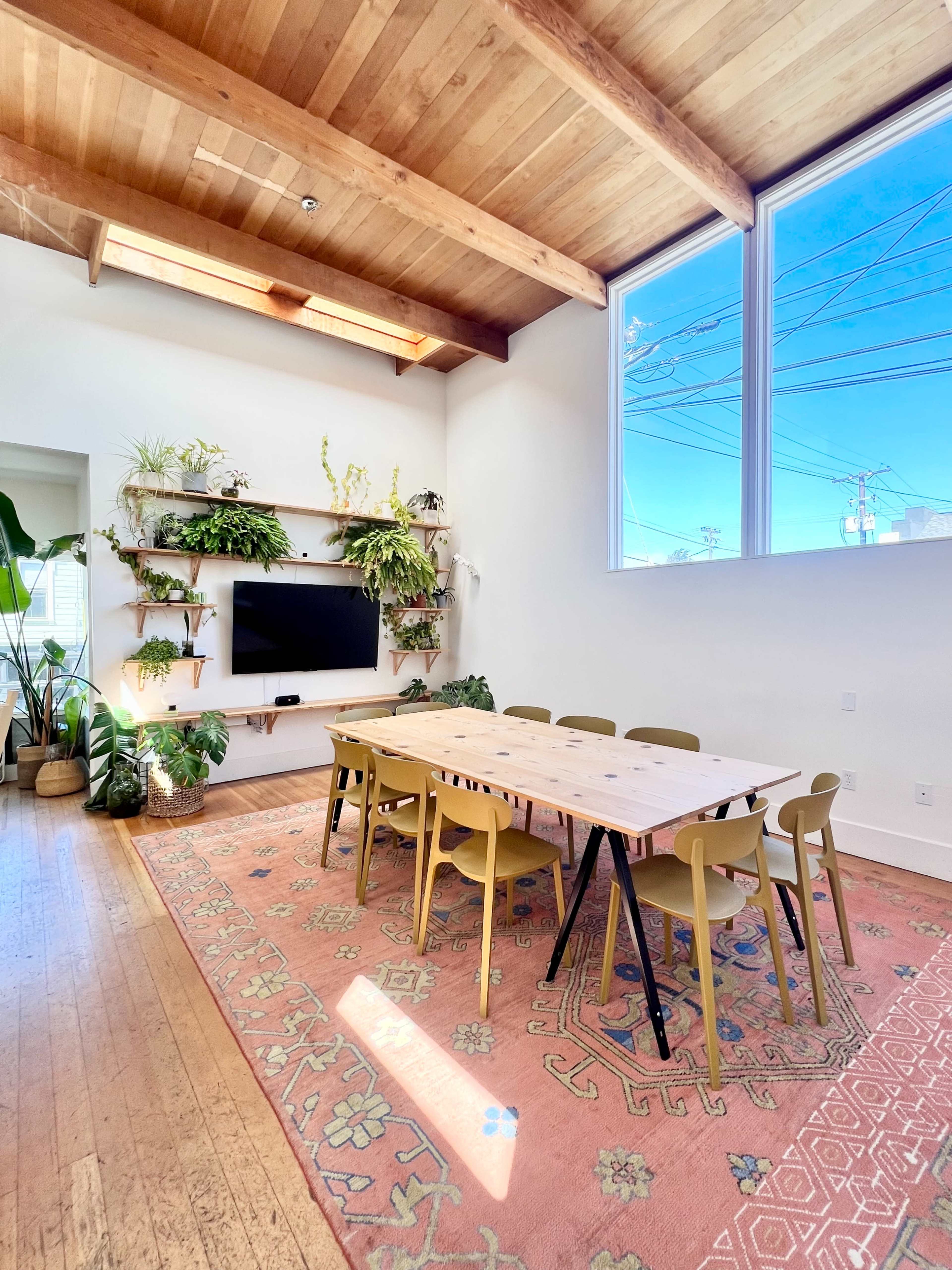The image shows a bright dining area with a wooden table surrounded by eight chairs, a rug on the floor, and plants adorning the walls and shelves.