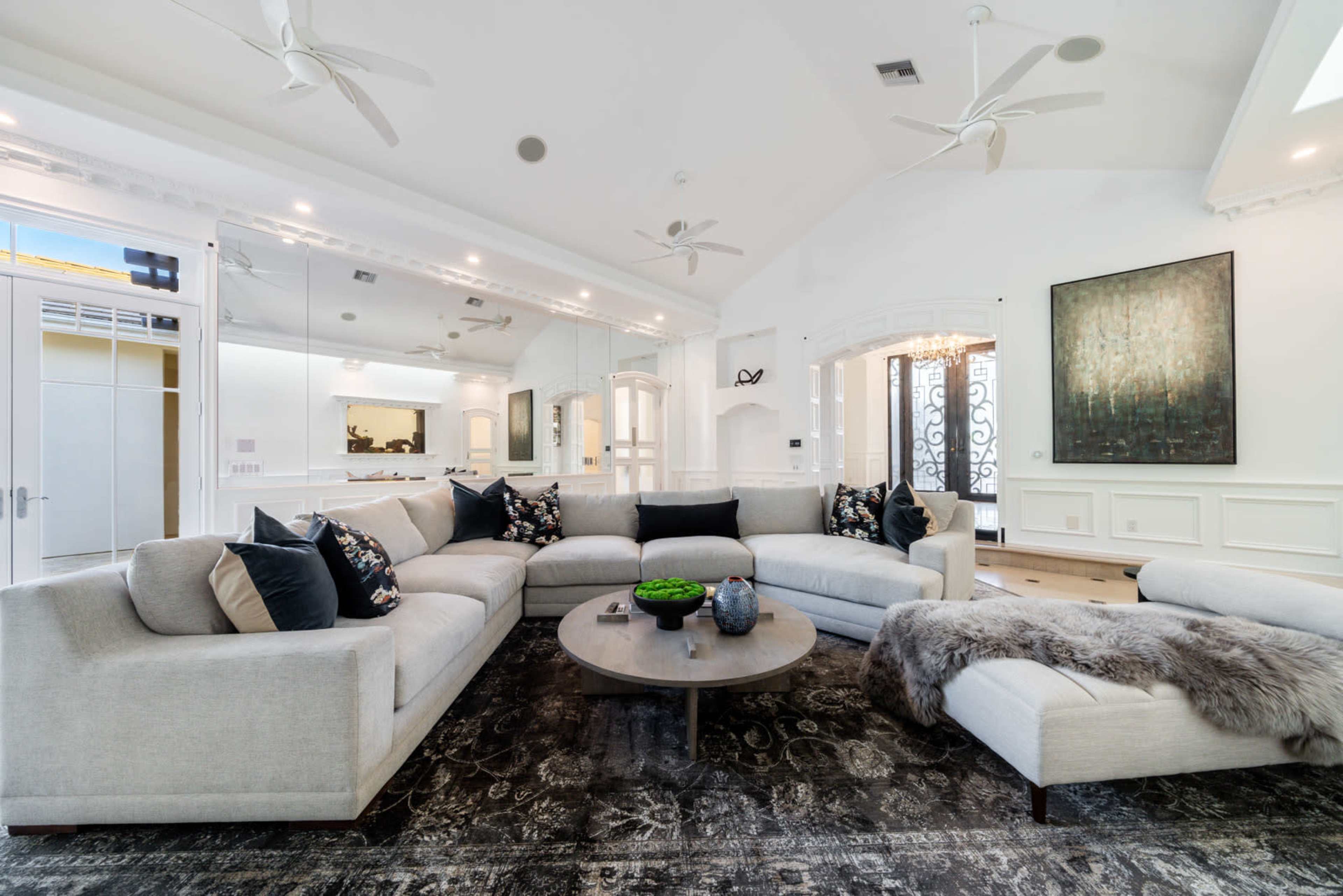 A modern living room features a spacious sectional sofa with decorative pillows, a round coffee table, and a large area rug, all set against white walls and bright lighting.