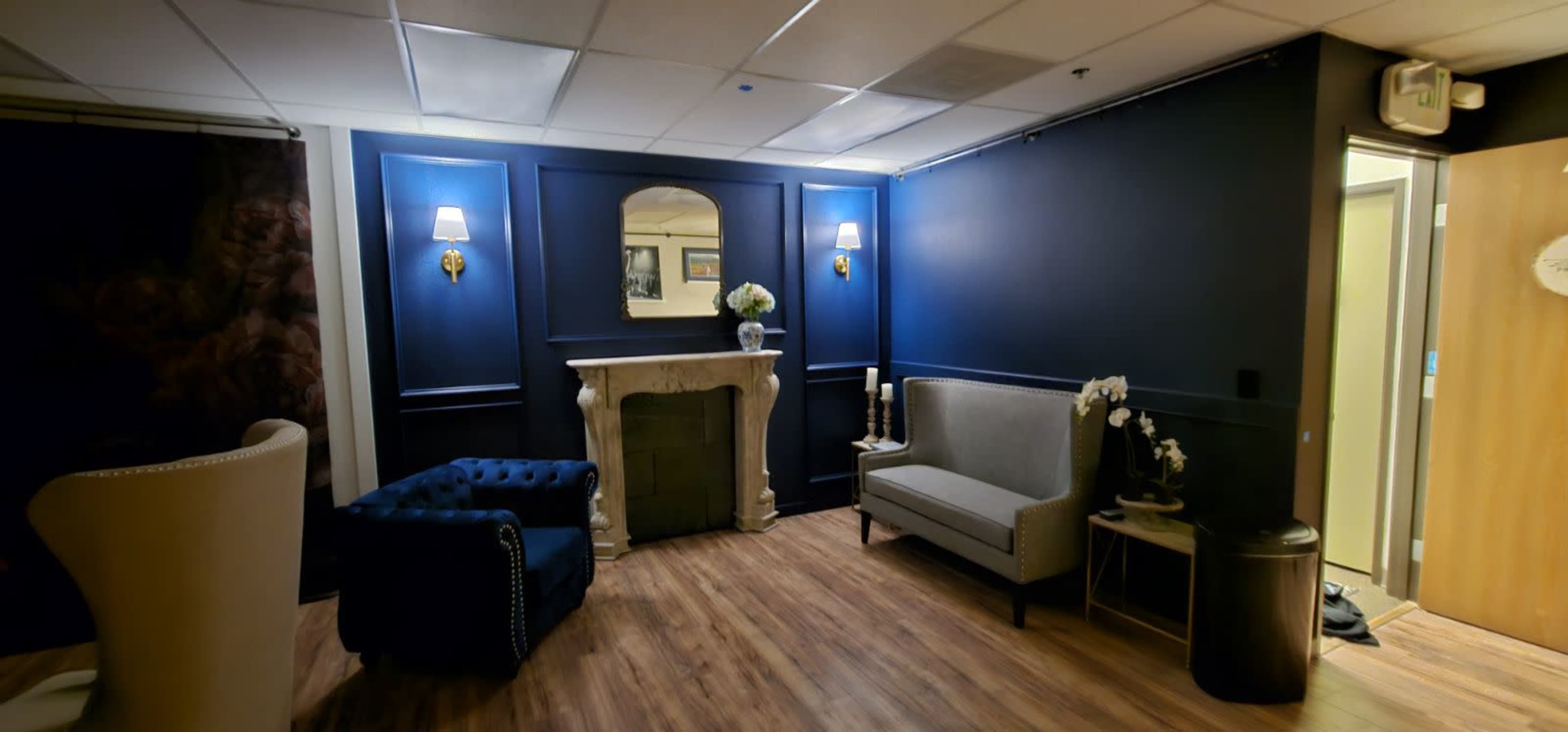 The image depicts a modern lounge area with dark blue walls, a decorative fireplace, a blue tufted armchair, a gray upholstered sofa, and a small table with floral decor.