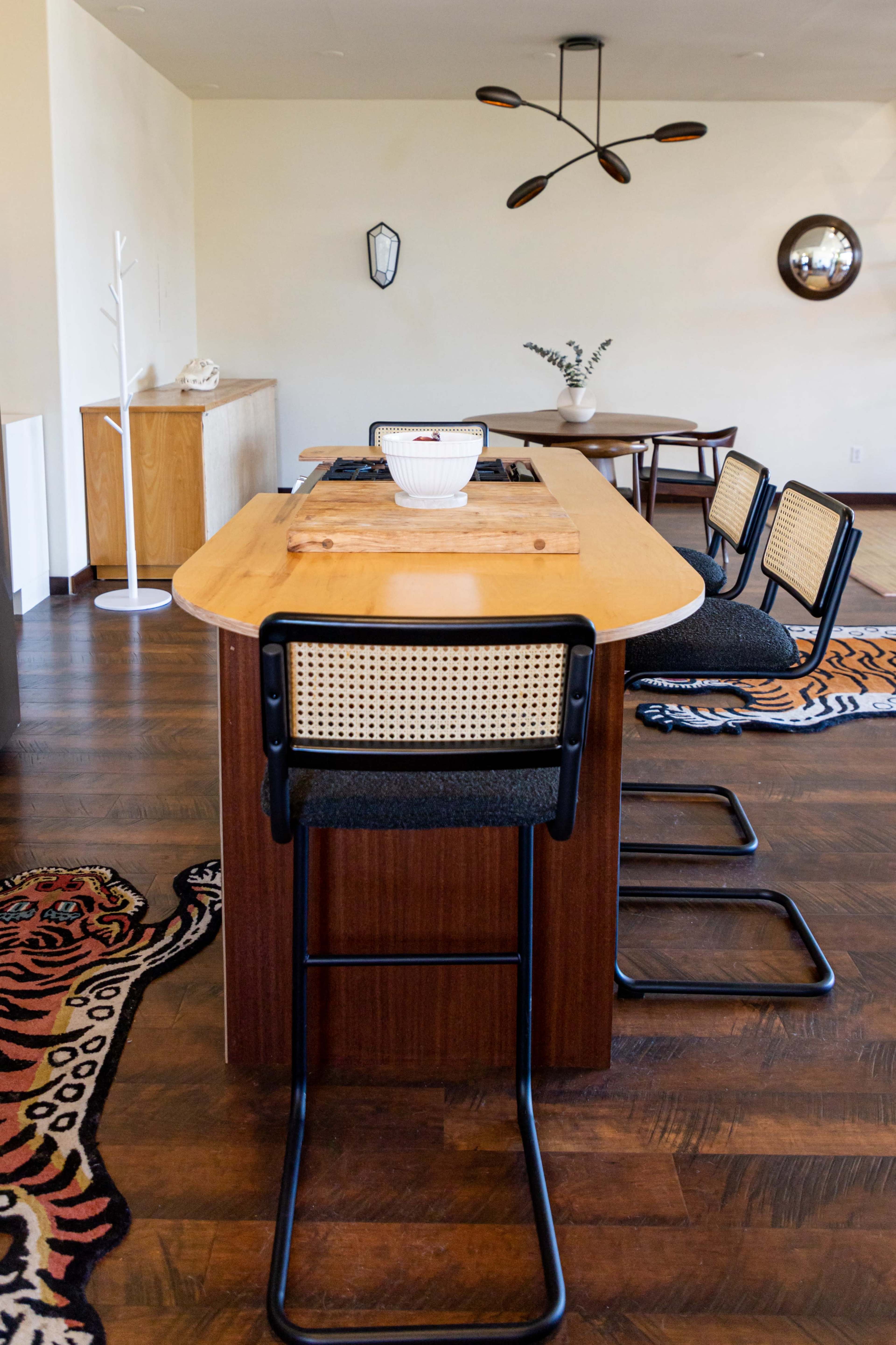 The image shows a modern kitchen dining area featuring a wooden island with three black and tan chairs, a decorative bowl on the island, and a rug with a tiger pattern on the floor.