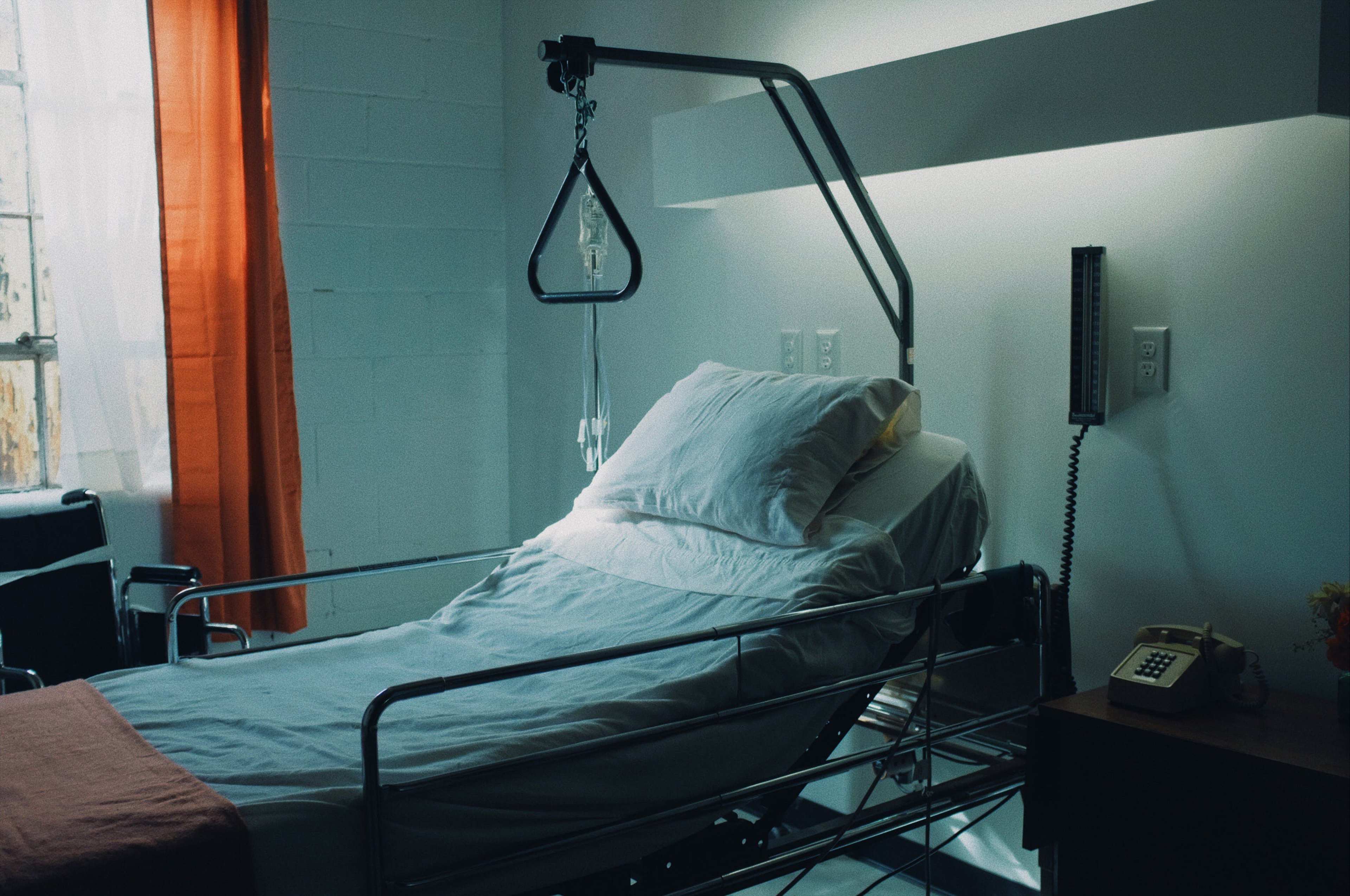 A hospital room features a metal hospital bed with white linens, a nightstand, and an IV stand next to a telephone.