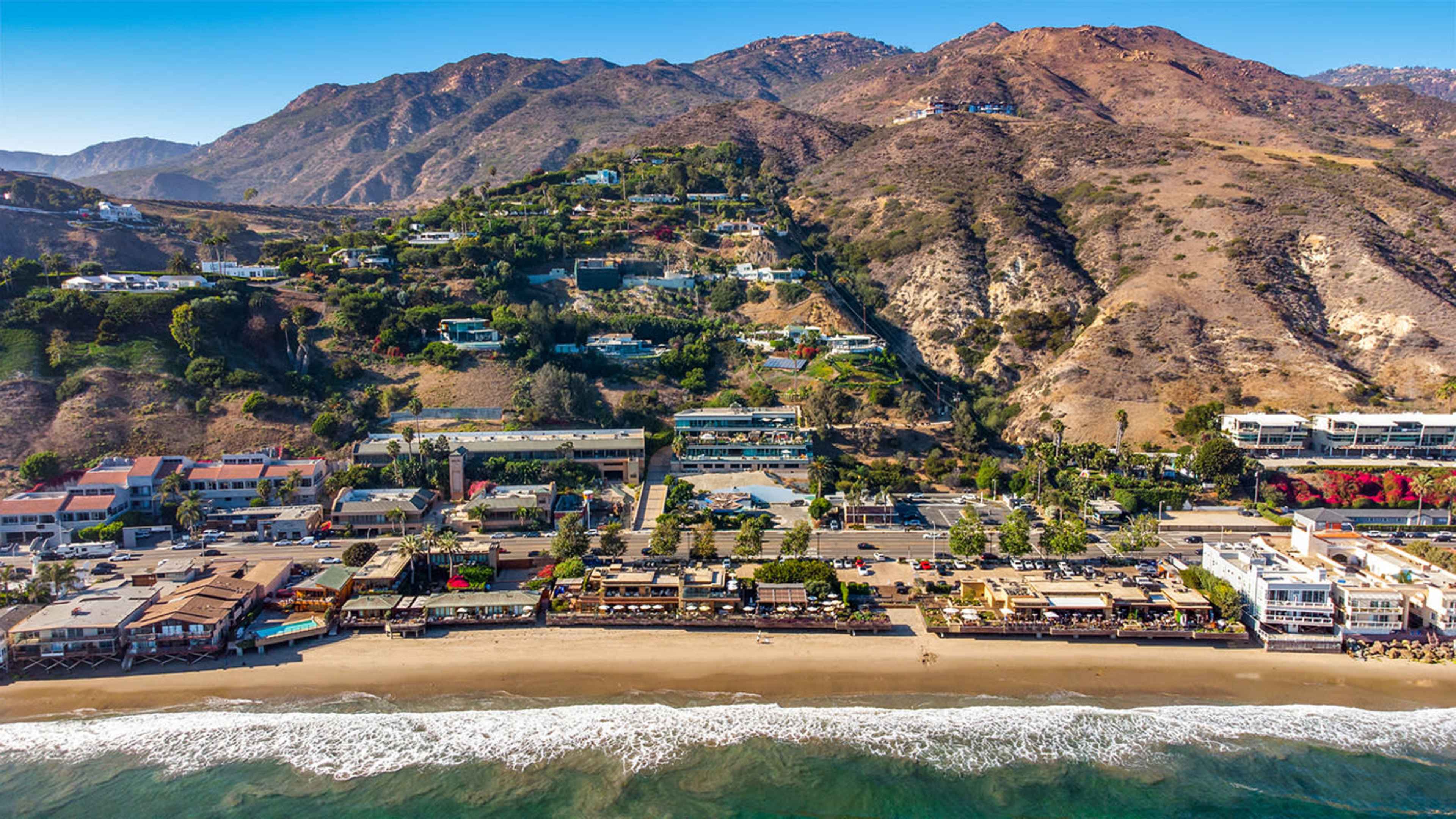 Aerial view of a coastal area featuring a sandy beach, a row of buildings, and hills in the background.