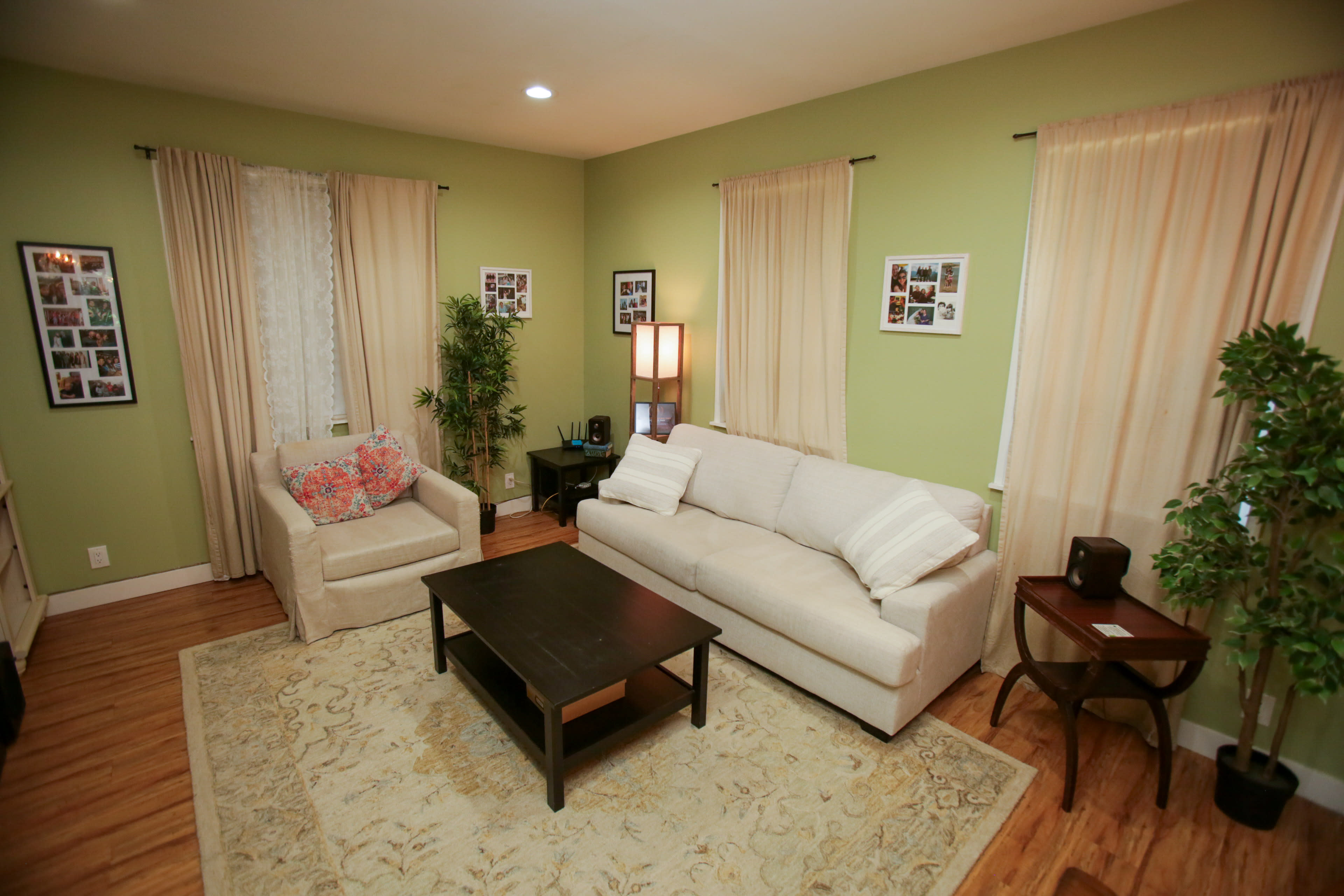 The image shows a cozy living room with a light-colored sofa, a beige armchair, a dark coffee table, and green walls adorned with framed photos.