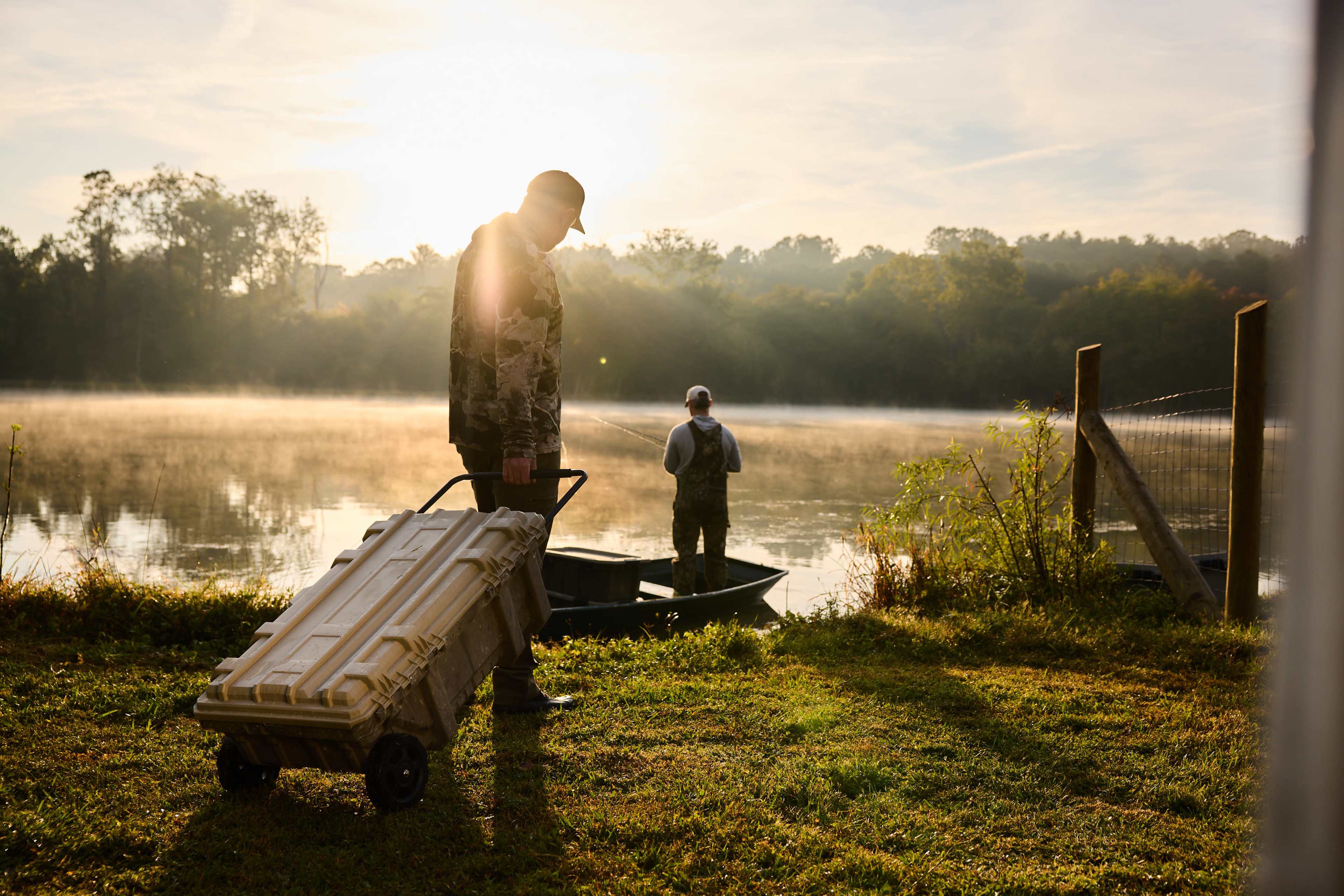 Two fishermen are by a misty river at sunrise, one standing with a cart and the other casting a line into the water.