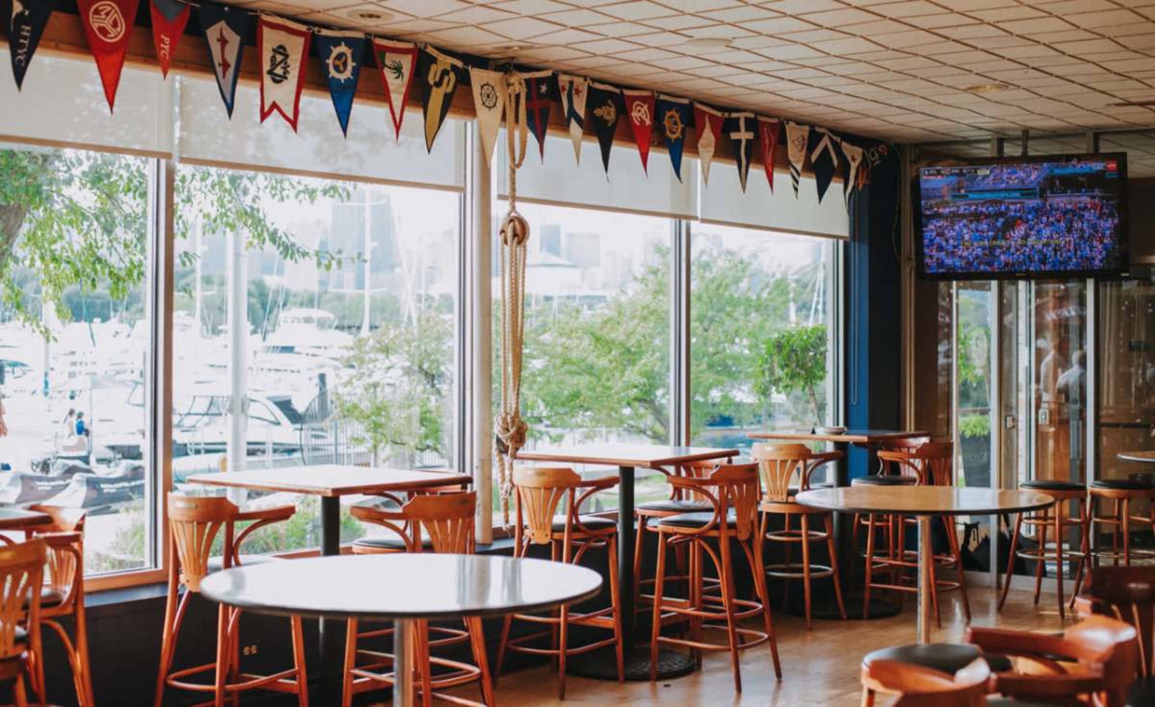The image shows a dining area with several wooden tables and chairs, large windows displaying a marina view, and a television mounted on the wall.